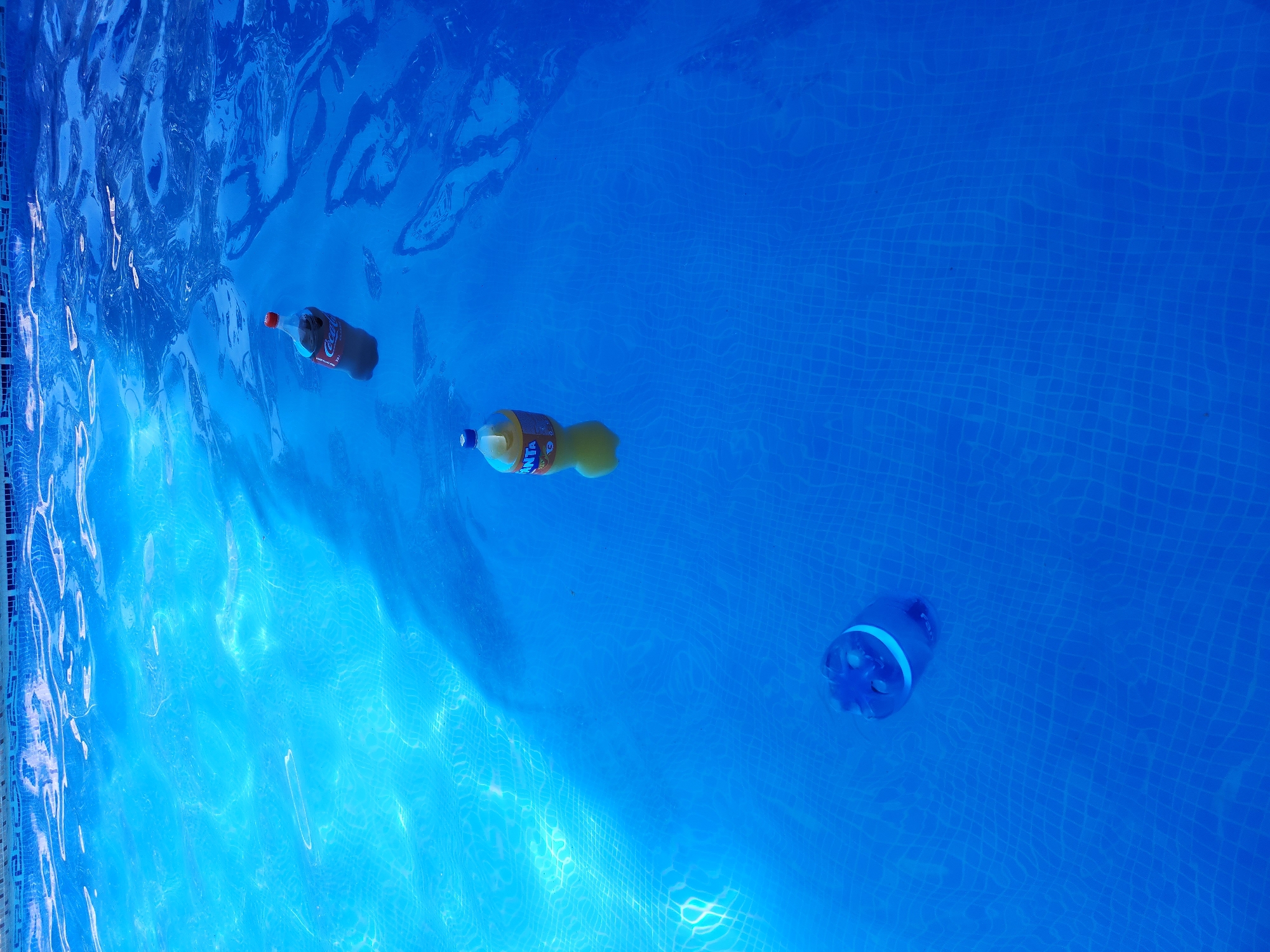 In the bright blue, sunlit waters of a swimming pool, three plastic bottles float. One bottle, a dark red Coca-Cola, is positioned near the top right of the frame, its cap a vibrant red. To its left and slightly lower, a bright orange Fanta bottle bobs gently on the surface, its label visible. At the bottom left of the image, a blue plastic bottle, possibly a water bottle, is partially submerged and tilted, its cap a lighter shade of blue. The water is clear, revealing the checkered pattern of the pool tiles beneath. The scene is devoid of human presence and any indication of its location within Yangikurgan, Uzbekistan.