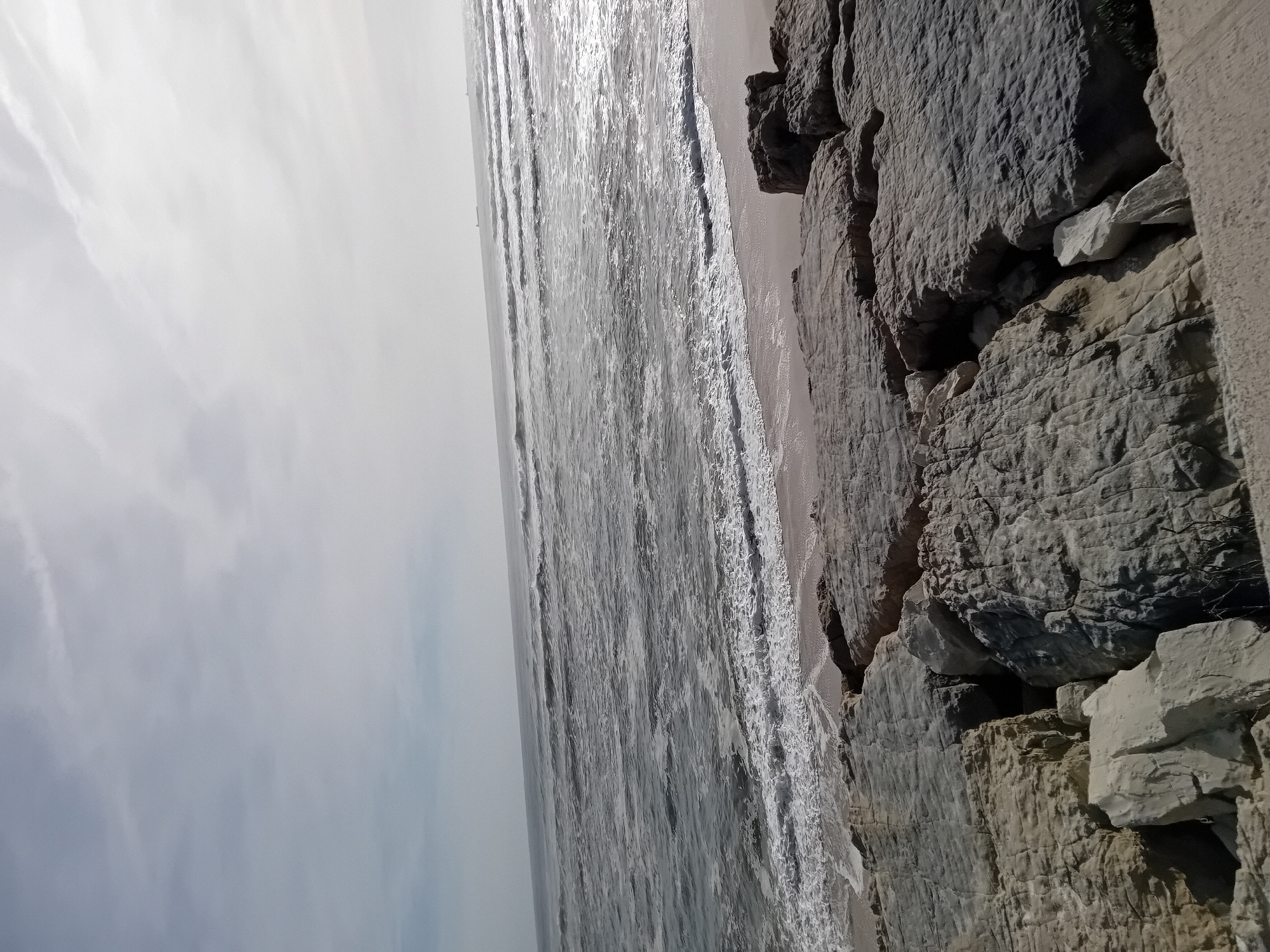 The image depicts a serene coastal landscape during daytime. The scene is dominated by a vast expanse of ocean, a sandy beach, and a rugged rocky cliff face.
The top portion of the frame shows a bright, somewhat overcast sky with subtle, wispy cloud formations, hinting at a bright, diffuse light source, likely the sun. Below the sky, the ocean stretches towards the horizon, displaying numerous waves breaking and receding towards the shore. The water's surface reflects a strong glare from the sun, particularly in the middle distance, creating shimmering streaks of silvery white.
A narrow strip of light-colored sand forms the beach, separating the ocean from a prominent rocky shoreline or cliff that occupies the right side and bottom of the frame. These rocks are predominantly dark grey and earthy brown, featuring deeply textured and layered surfaces. Some sections of the rock appear lighter, almost beige or off-white, and there are visible crevices and shadows indicating the rough, uneven terrain.
No people, man-made objects, or visible text are present in the image. The time of day appears to be mid-day, given the bright, overhead lighting and the strong reflections on the water. The weather seems clear to partly cloudy. The scene conveys a sense of natural, untamed beauty and coastal tranquility.