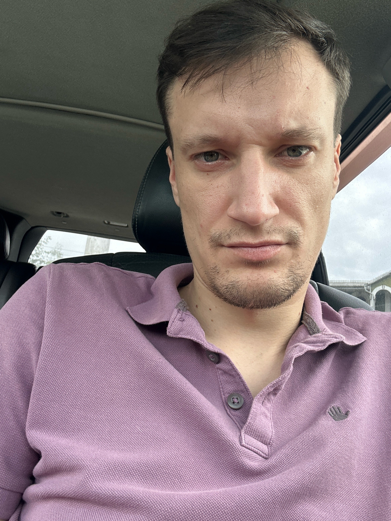 A man with short brown hair and a light beard is shown from the chest up, sitting in the front seat of a car. He is wearing a purple polo shirt. The interior of the car is visible behind him, including the headrest of the seat. The exterior of the car is seen through the window, showing a building with a patterned facade. The man's expression is neutral. This image was taken in Shatava, Ukraine.