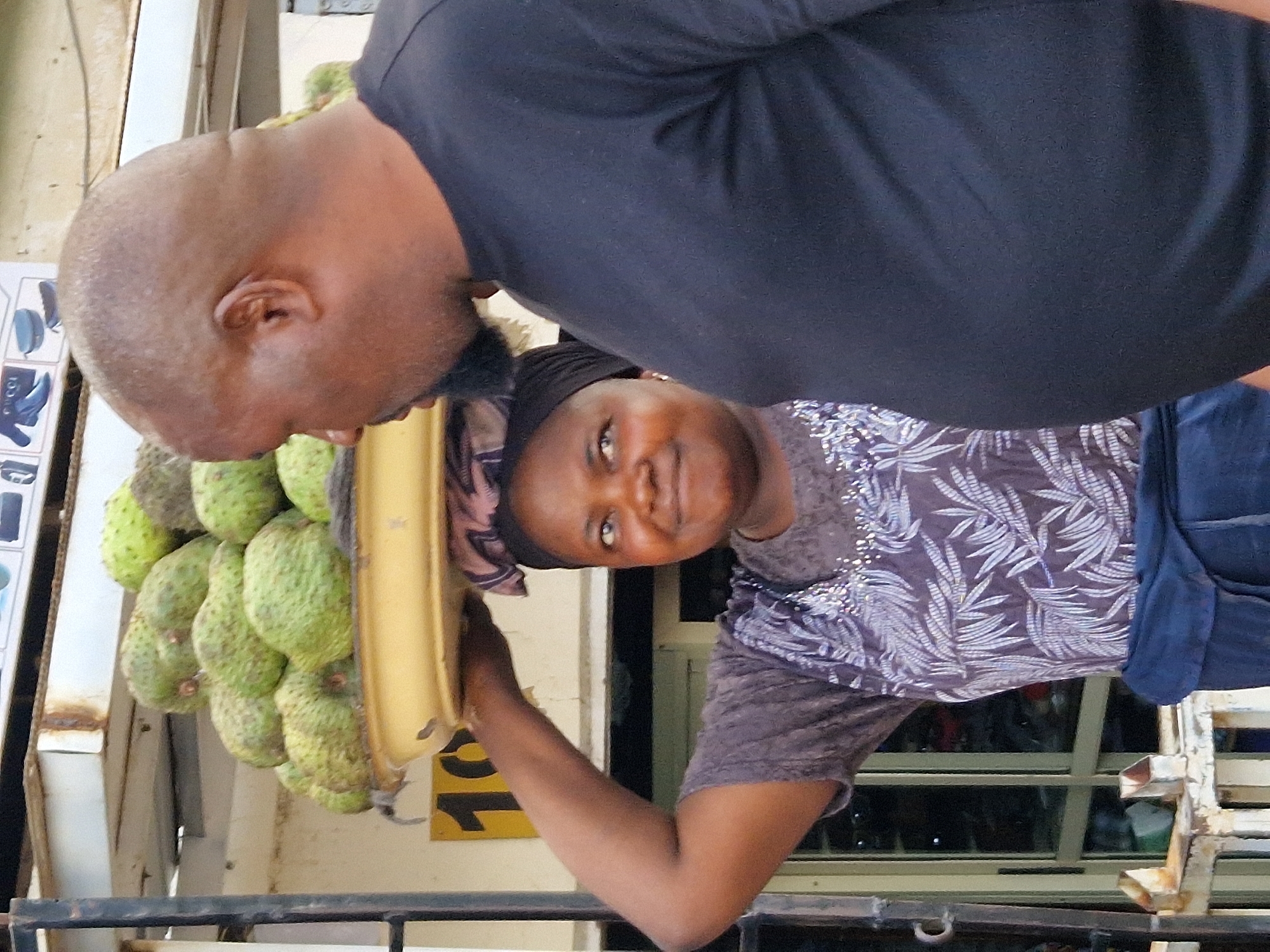 In an outdoor setting in Unknown, Unknown, a Black woman balances a large tray of soursop fruit on her head, her right arm raised to support it. She wears a dark headscarf, a patterned grey and white t-shirt, and a blue wrap around her waist. To her right, a Black man with a shaved head and a goatee stands close, wearing a black t-shirt. The background shows a stall with metal shelving and a sign with the number "10" on it.