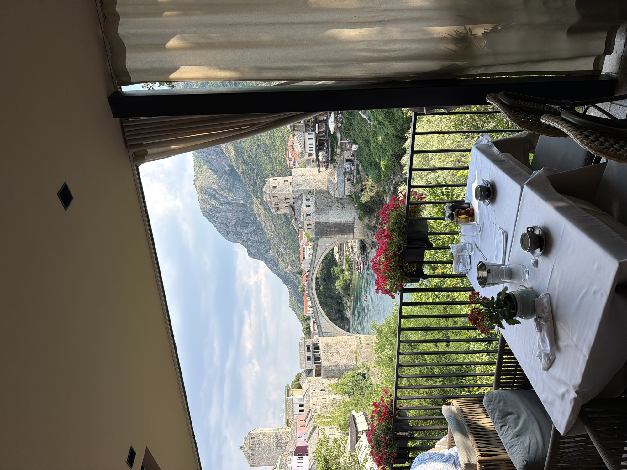 The image displays a scenic view from a restaurant balcony overlooking a river and an iconic bridge. In the foreground, a table is set for dining with white tablecloths, cups, saucers, and what appear to be salt and pepper shakers, and a water pitcher. Wicker chairs with cushions are visible, suggesting a comfortable outdoor dining area. Flower pots with red flowers adorn the railing of the balcony, adding a touch of color.
The background features a historic stone bridge arching over a turquoise river. Beyond the bridge, traditional stone buildings cascade down a hillside, and a large, imposing mountain rises dramatically against a bright blue sky with scattered clouds. The sunlight suggests it is daytime, likely late morning or afternoon.
The scene evokes a sense of relaxation and enjoyment, with the beautiful natural landscape and the historic architecture creating a picturesque setting. There is no visible text in the image.