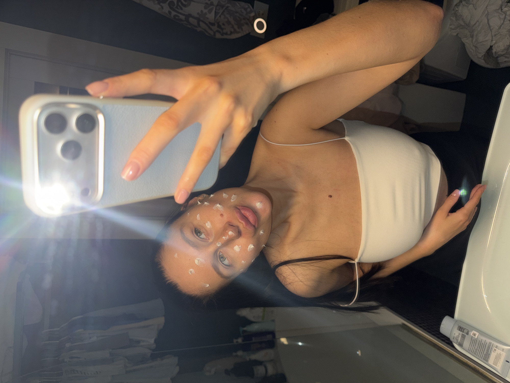 This image captures a mirror selfie of a young woman in an indoor setting, likely a bathroom or bedroom, given the presence of a sink and what appears to be a bed or clutter in the reflection. The image is oriented sideways, with the woman looking towards the camera of her smartphone.
The primary subject is a woman with dark hair, visible light-colored eyes, and a neutral expression. Her face is dotted with numerous small, white spots of what appears to be a topical skincare product, such as an acne treatment or spot cream, covering her forehead, cheeks, nose, and chin. She is wearing a white, ribbed camisole or tank top with thin spaghetti straps.
She is holding a light-colored (possibly light blue or grey) smartphone, which appears to be an iPhone model with three camera lenses. The phone's flash is active, creating a bright, circular flare of light on the left side of the image, illuminating her face and the immediate surroundings.
To the right, a white sink basin and a dark countertop are partially visible. A light grey tube, possibly containing another skincare product, rests on the counter with some illegible text on its label. The background, largely obscured by the mirror's reflection and the dark room, shows hints of patterned fabric (like bedding) and dark furniture or shelving. The overall lighting suggests a dim environment, enhanced by the phone's flash. The location context for this image is Khimki, Russia.