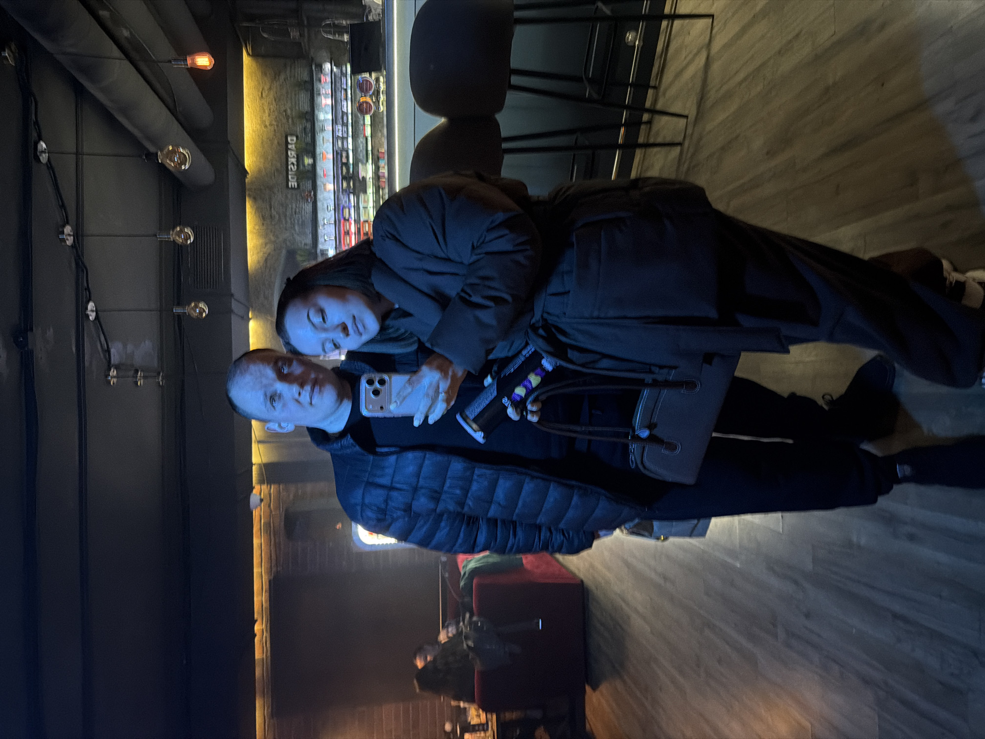 The image captures two people, a man and a woman, posing for a photo in what appears to be an indoor bar or lounge setting during the evening or night. The man, on the left side of the frame, is in the foreground, wearing a dark puffer jacket. He is holding a smartphone with a white case, likely taking a selfie, and is looking directly at the camera. The woman is positioned behind and to the right of the man, resting her head gently on his back or shoulder, with her eyes partially closed. She is dressed in a dark, long coat or jacket and carries a dark handbag over her arm.
The background reveals a dimly lit but inviting atmosphere. Behind the couple, there is a bar counter with various bottles and items displayed on shelves. Above the bar, a prominent sign with the text "DVЯKSIDE" (likely "DARKSIDE," using a stylized 'Я' for 'R') is visible. The decor includes industrial-style elements such as exposed pipes and multiple warm, incandescent light bulbs hanging from the ceiling and strung along wires, providing ambient lighting. A section of a brick wall and wooden flooring can also be seen. Further in the background, out of focus, a red couch and vague figures of other patrons suggest a busy but relaxed environment. The overall impression is one of a casual, cozy establishment.
