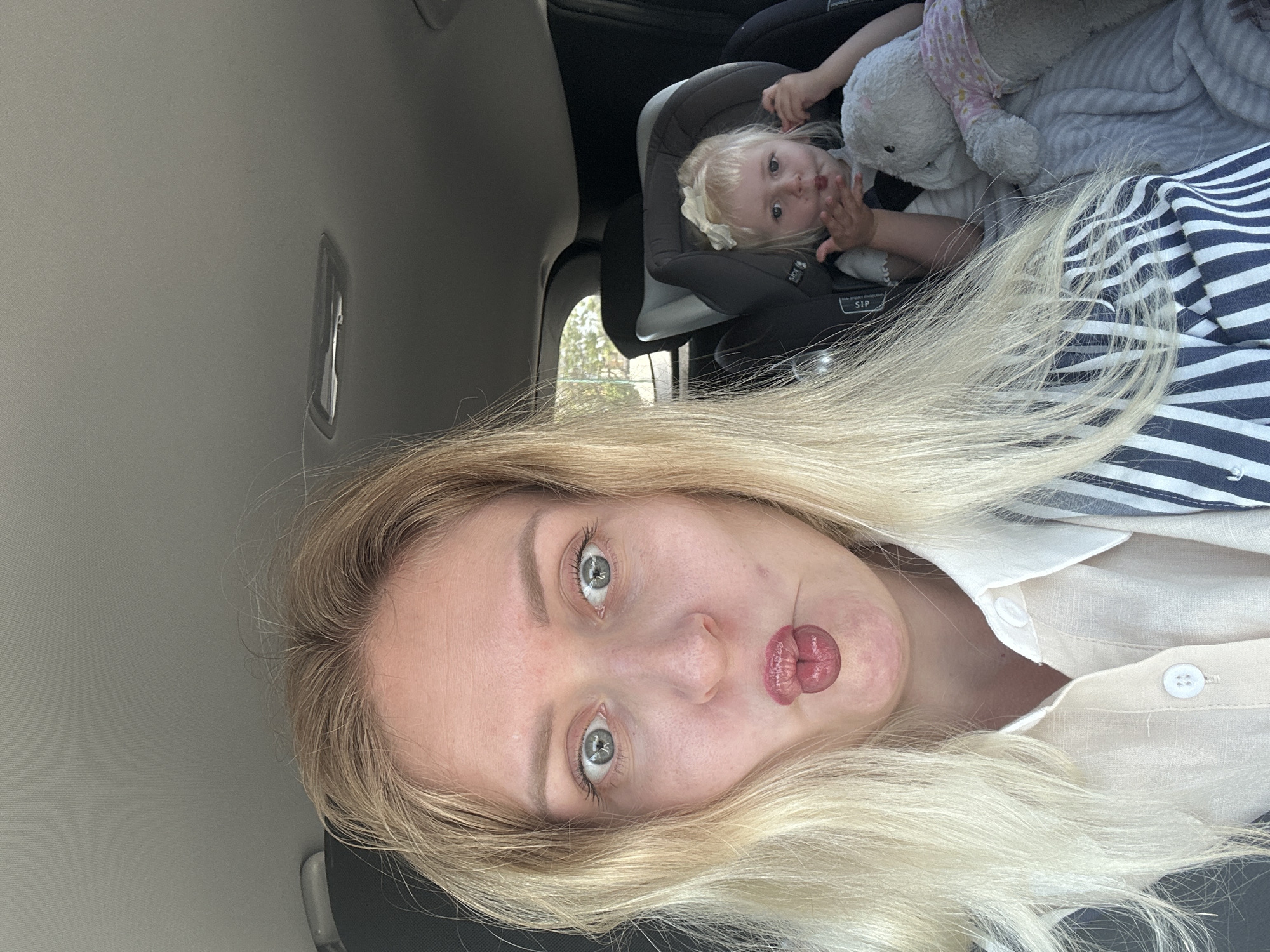 A selfie taken inside a vehicle. A blonde woman with exaggerated puckered lips is in the foreground. Behind her, a young child sits in a car seat with a stuffed animal. The car's interior is visible. The location is unknown, and the request to specify Petah Tikva, Israel, cannot be substantiated from the visual information provided.