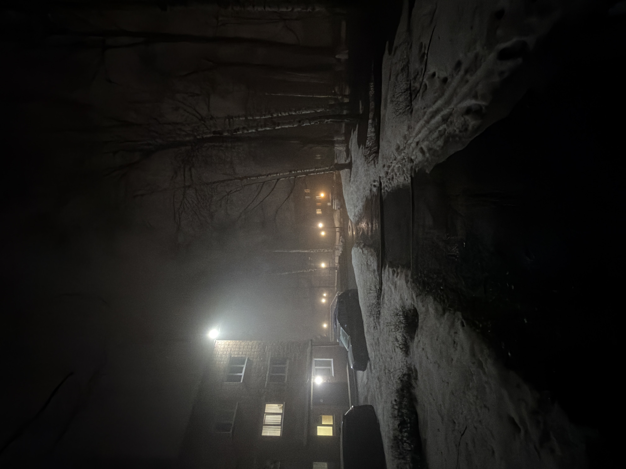 A night scene in Sarapul, Russia, shows a dark, possibly snowy or icy street illuminated by streetlights and building lights. A three-story brick building with multiple visible windows, some illuminated, is seen on the left. The street appears to be lined with snow-covered embankments and potentially parked vehicles. Bare trees are visible in the background, obscured by fog or mist. The lighting suggests a cold, urban environment.