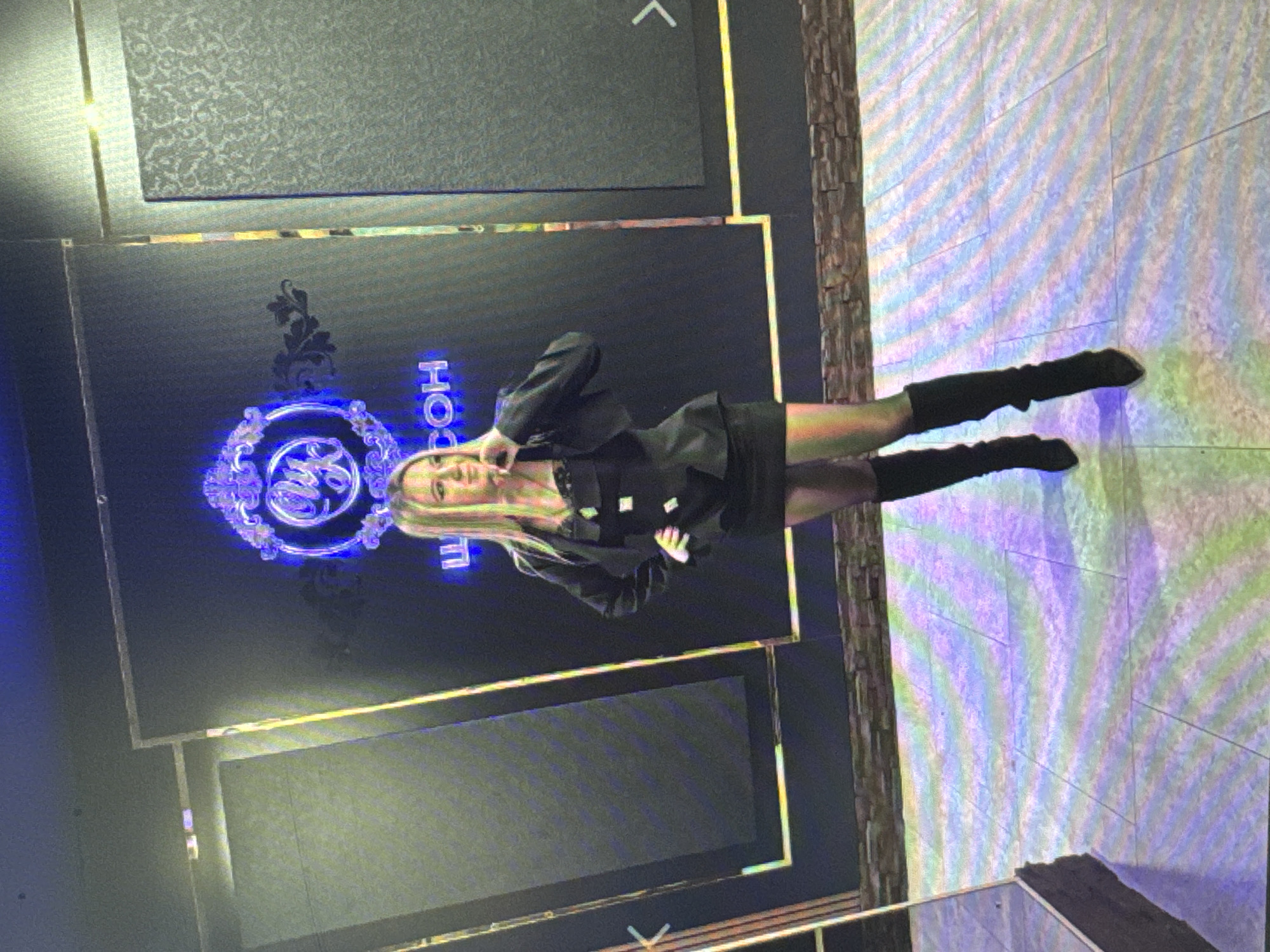 An adult female stands in an indoor environment, posing with her right hand touching her chin. She is wearing a black blazer over a black dress, black thigh-high boots, and sheer black stockings. A large, illuminated logo with intricate detailing is visible behind her. The logo is framed by a purple neon light and appears to be in Cyrillic script, with the visible portion reading "COH". The background is dark, with what appear to be textured panels on either side of the central logo display. The floor is tiled and reflects a colorful, iridescent pattern. The overall lighting is dim, with accent lighting from the logo and the floor. The scene suggests an interior space, possibly a commercial establishment or event venue.