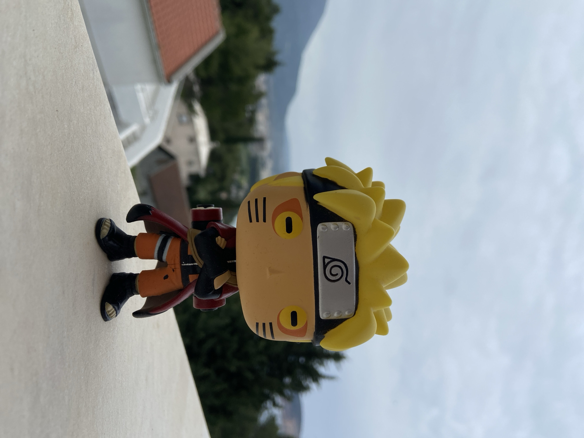 A Naruto Uzumaki Funko Pop figurine stands on a ledge, likely a balcony or wall, overlooking Šušanj, Montenegro. The backdrop features buildings, trees, and mountains under an overcast sky. The figurine is wearing its signature orange and black outfit, and the backdrop gives a sense of a residential area nestled within a mountainous region.