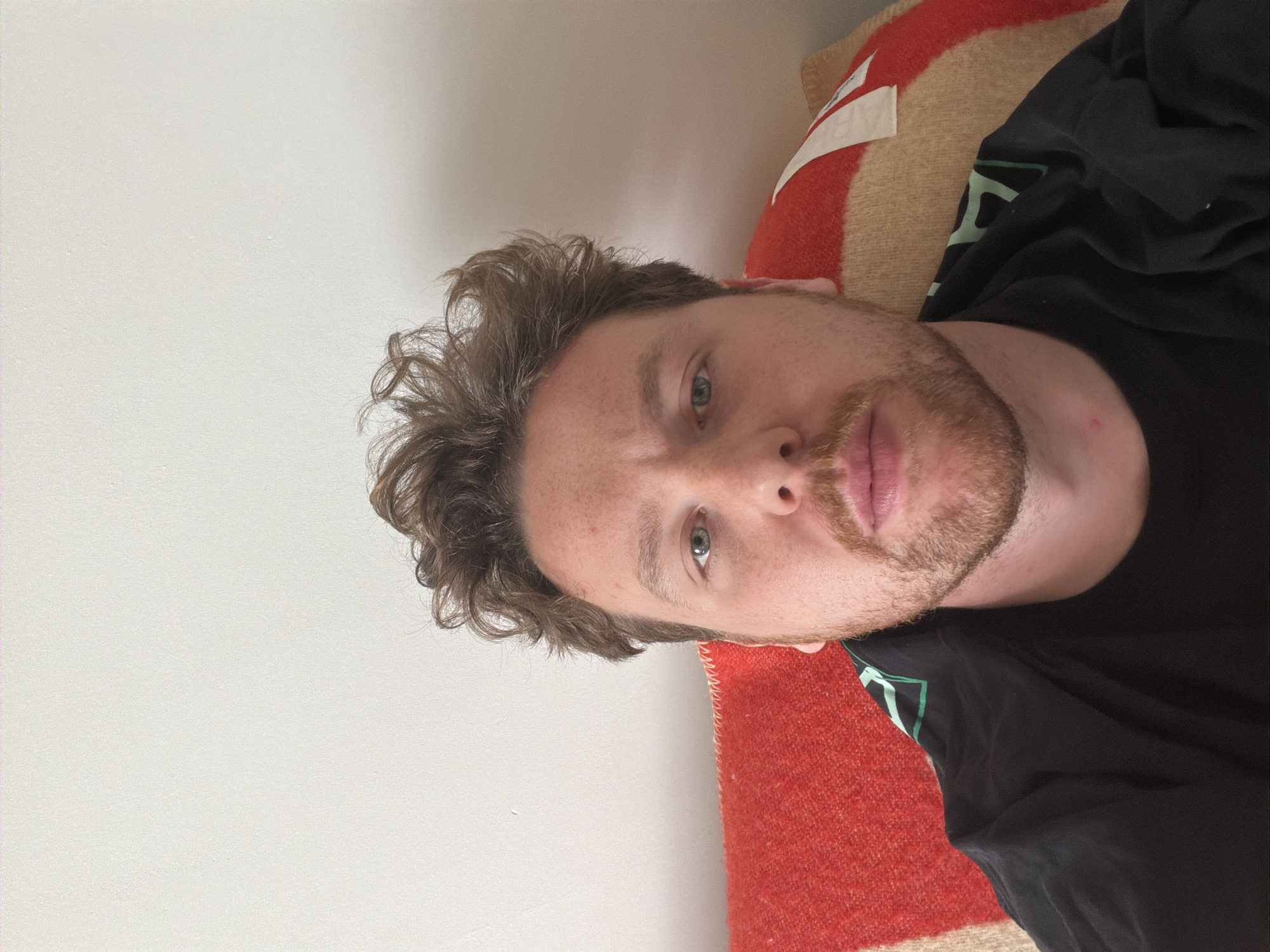 A male subject is depicted in a close-up, eye-level indoor portrait. The scene is set within an interior room in London, United Kingdom. The subject has fair skin, light green eyes, and light brown, wavy, disheveled hair, and a light brown beard and mustache. He wears a dark, possibly black, crew-neck shirt with a subtle lighter pattern on the left shoulder. Behind him, a textured blanket or cushion is visible, displaying horizontal stripes of red, light beige, and a thin white stripe. A small white tag is present on the beige stripe. The background above the subject and blanket is a plain, light-colored wall.