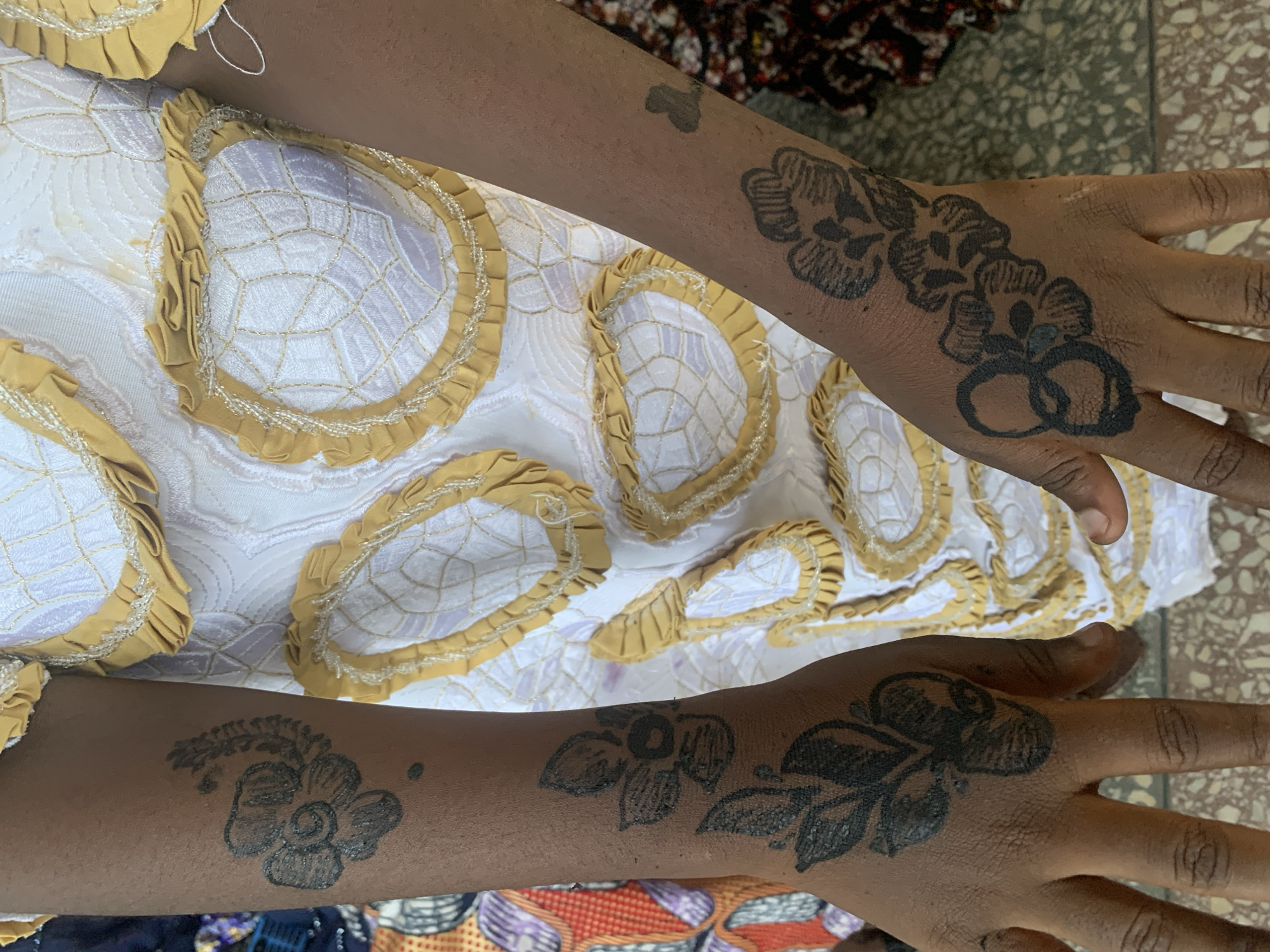 Two dark-skinned arms with henna tattoos are positioned over a white fabric with gold detailing. The tattoos depict floral and interconnected circle designs. The location is presumed to be Jos, Nigeria, based on the prompt.