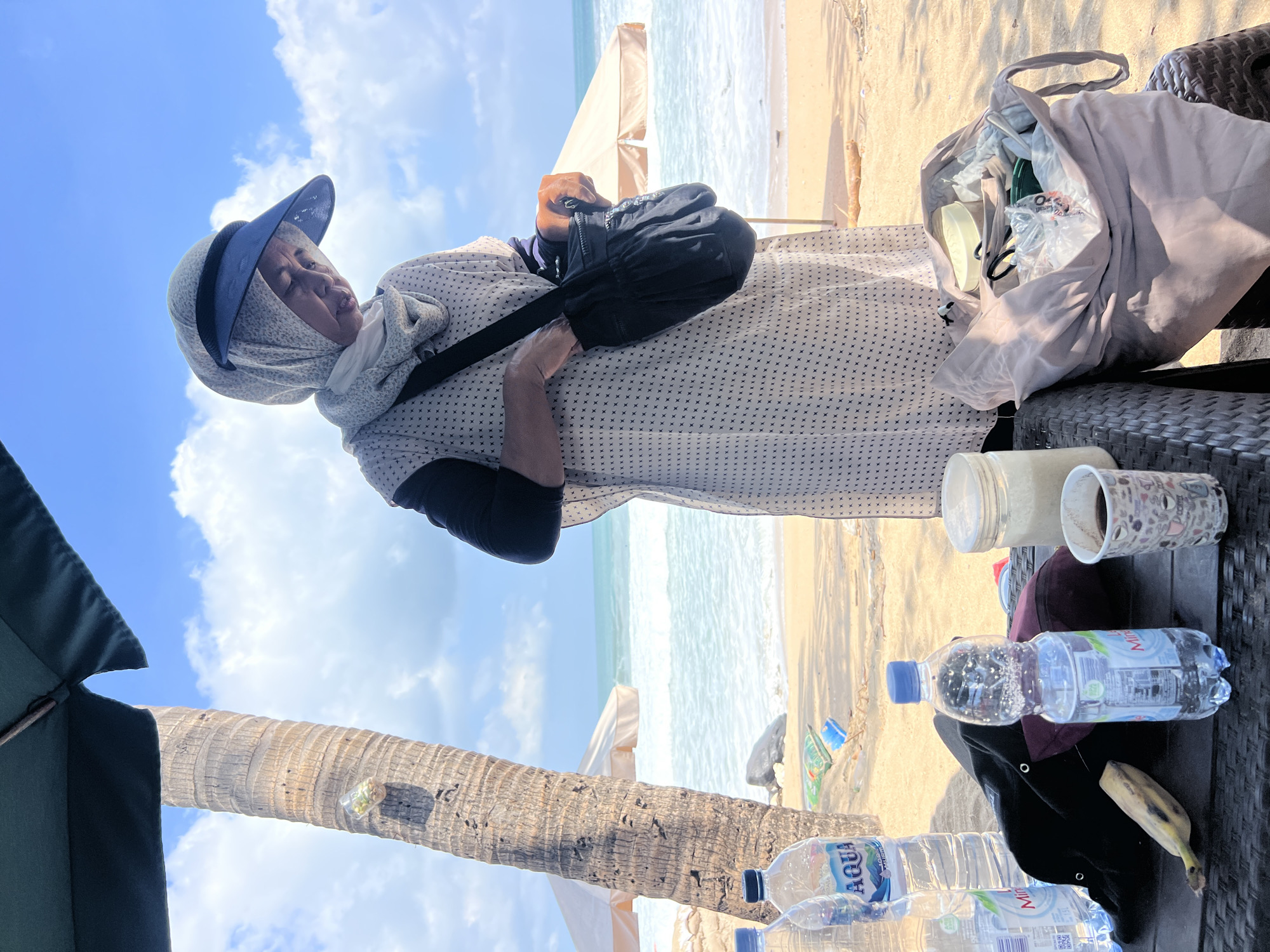 A woman wearing a hijab and a blue sun visor stands on a sandy beach in Kuta, Indonesia, holding a black bag. She is wearing a light-colored, patterned dress over a black long-sleeved shirt. In front of her, on a woven table, are two bottles of Aqua brand water, a banana, a cup with a coffee-like beverage, and a jar of sugar or similar granular substance. A large tote bag is open on a chair to her right, filled with various items. The ocean with moderate waves is visible in the background, along with a palm tree and beach umbrellas. The sky is blue with scattered white clouds.