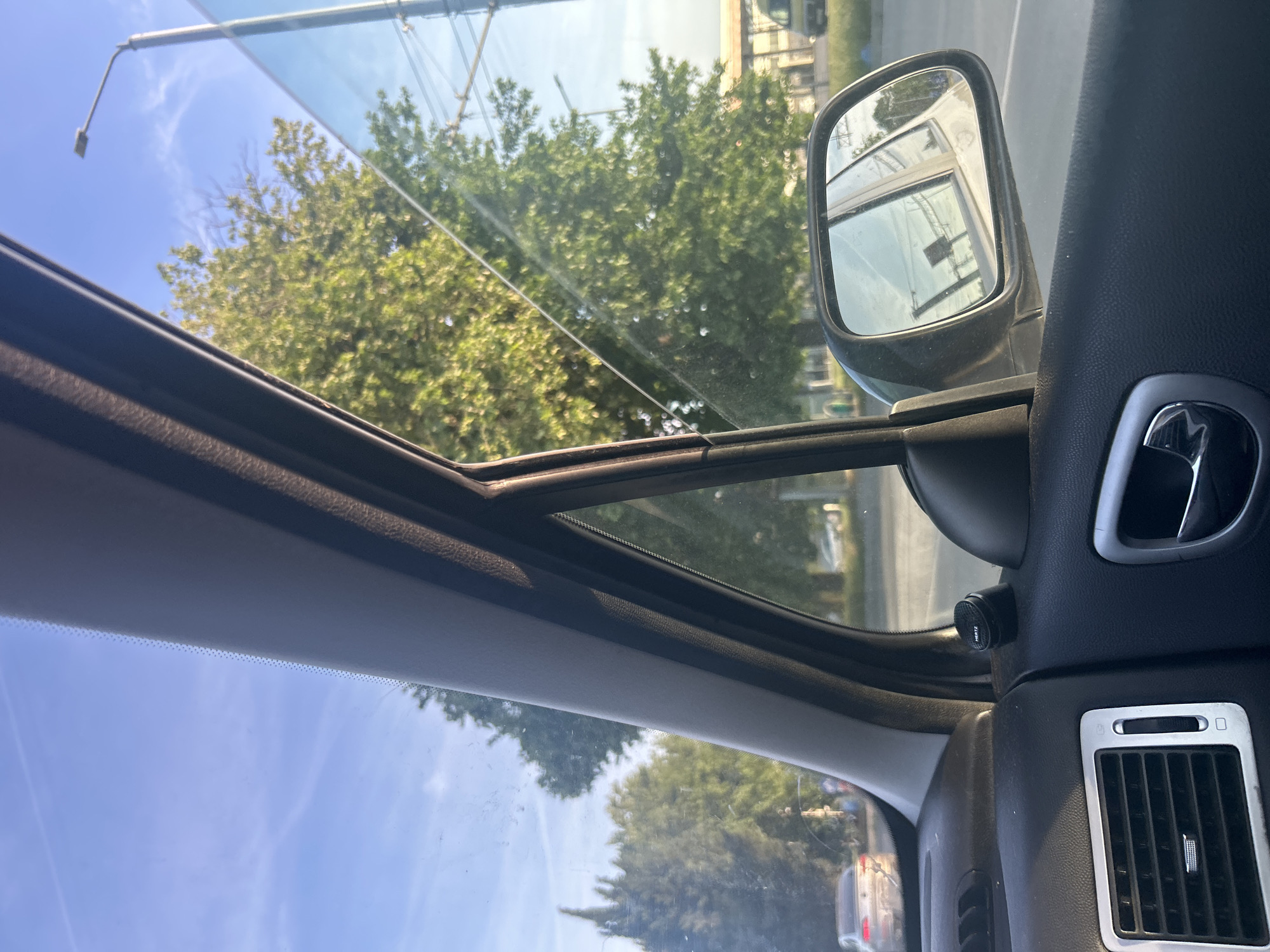 View from inside a car, looking out the windshield at trees and a blue sky. A car side mirror is visible on the right. The car's dashboard and air vents are also visible. The location is Unknown, Unknown.