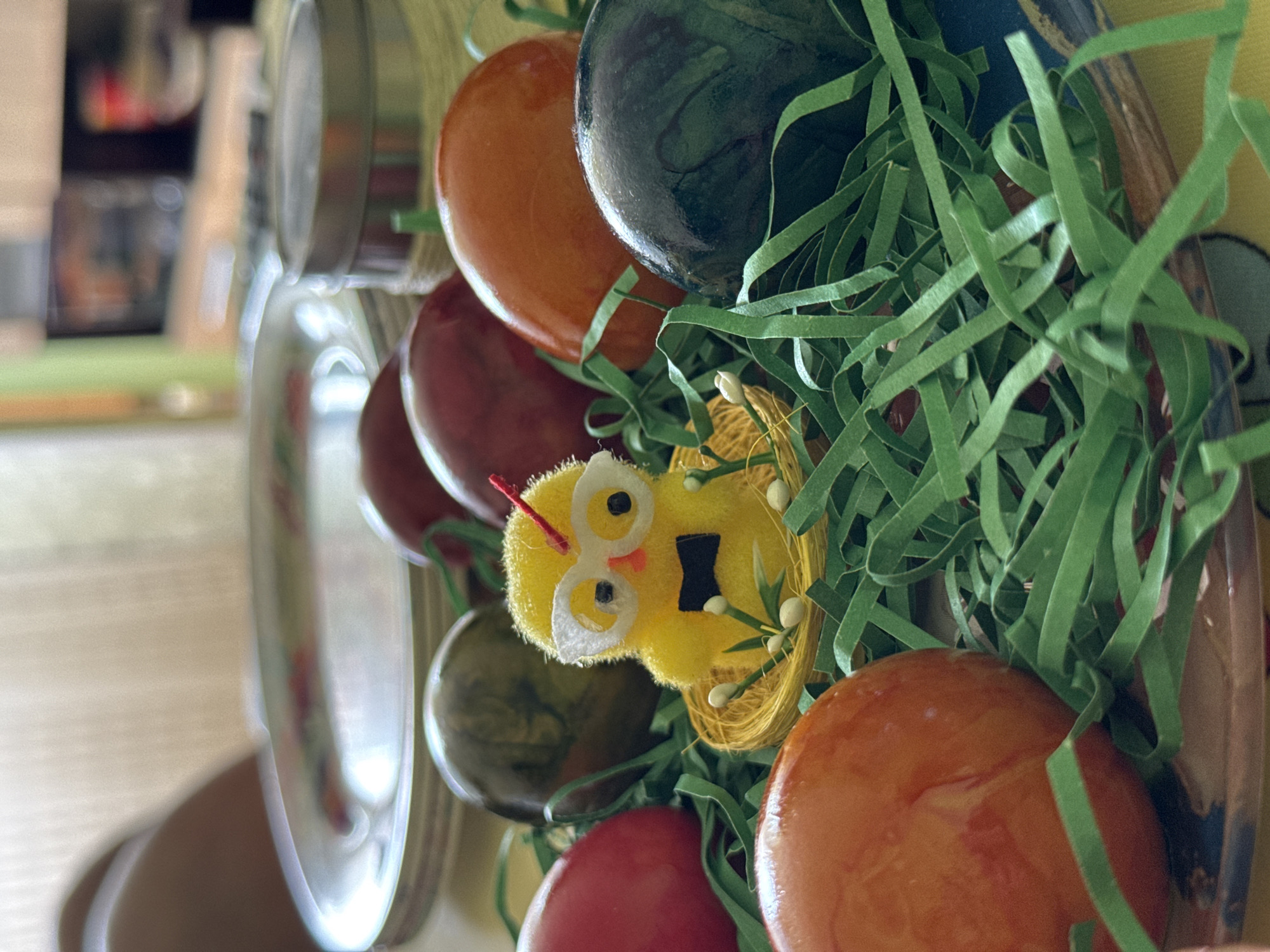 The image depicts a decorative arrangement of artificial Easter eggs and green paper grass, accented by a yellow chick ornament. The eggs are various colors including orange, red, and green, and appear to be made of a smooth, possibly glazed, material. The chick is yellow and has a small black cutout resembling a beak or mouth. The background is blurred, suggesting an interior setting with metallic containers visible in the periphery. There is no specific information within the image to determine that the location is in Burgas, Bulgaria. The scene likely represents an Easter-themed display or decoration.
