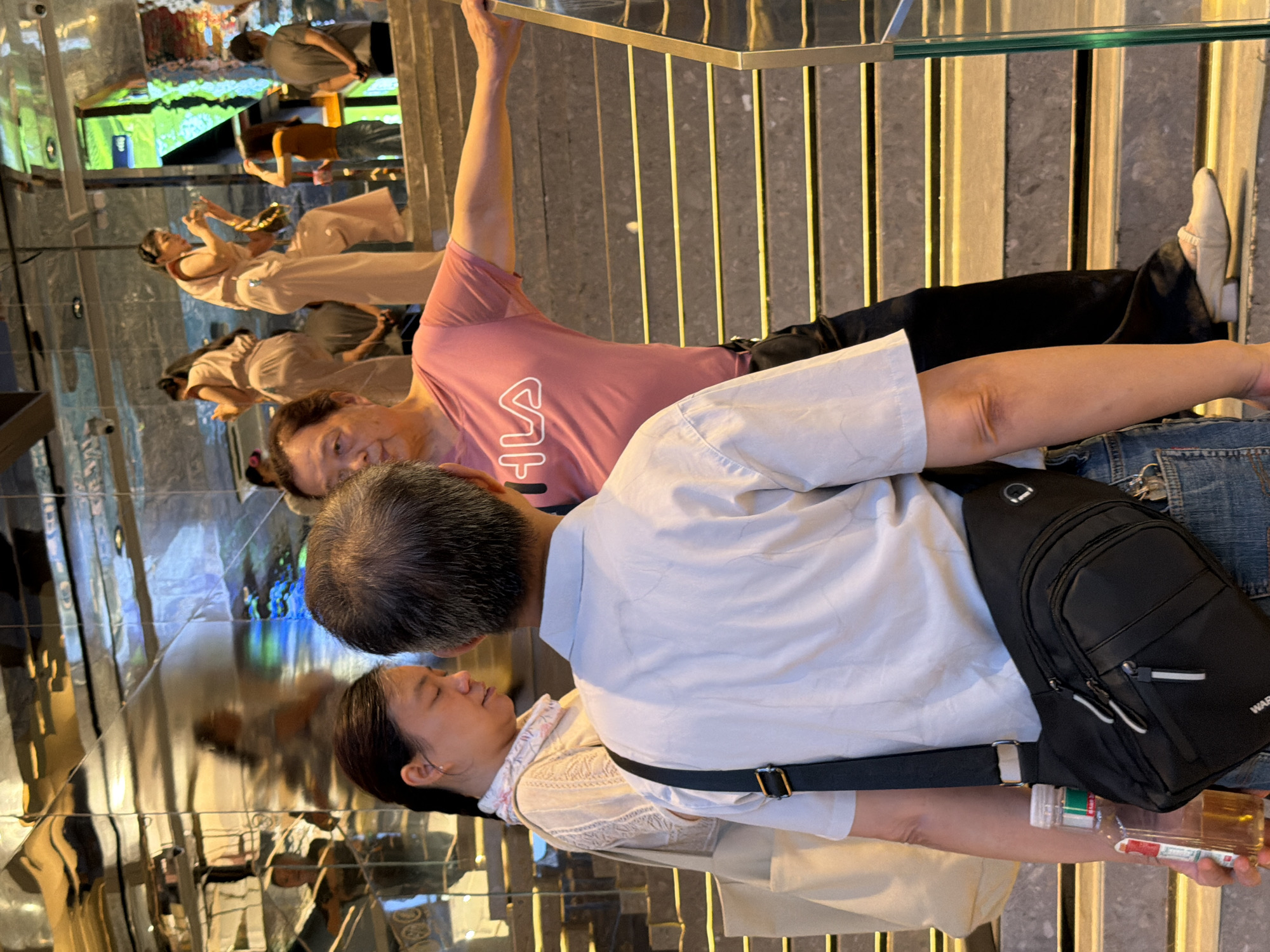 In Yuzhong District, China, three people are on a reflective, multi-level staircase. The person in the foreground, facing away from the camera, wears a light blue collared shirt and has a black crossbody bag. Next to them, a woman with dark hair pulled back is looking down, wearing a cream-colored textured top with a patterned scarf. To her right, a man in a pink t-shirt with a white "FILA" logo is leaning on the railing and looking towards the woman. In the background, several other individuals are visible on the stairs, some wearing light pink outfits. The staircase has yellow accents on each step and is bordered by a glass railing on the right. The environment has highly reflective walls and ceilings.