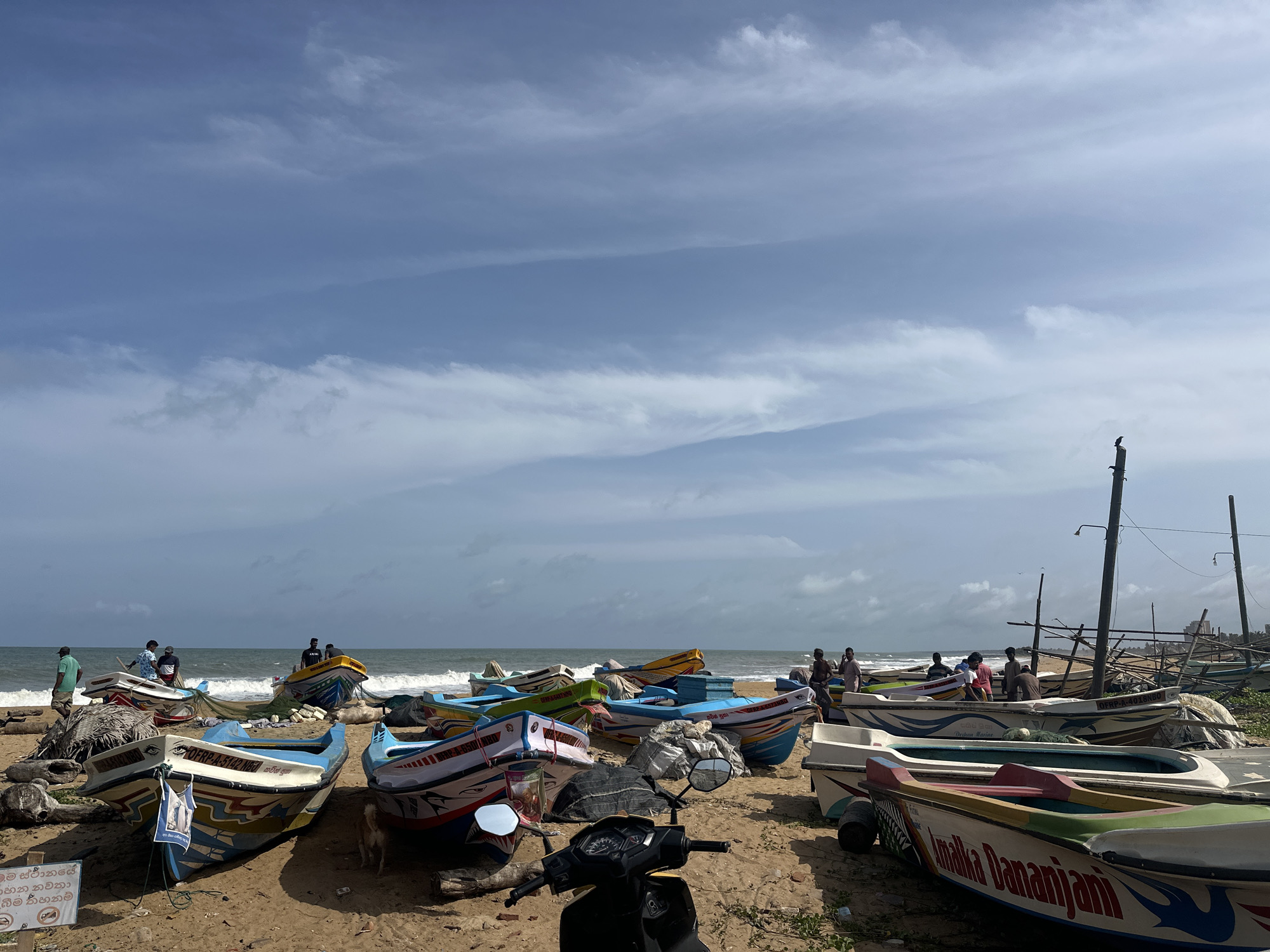 The image depicts a beach scene in Negombo, Sri Lanka, with numerous colorful fishing boats beached on the sand. The boats are of various sizes and designs, many adorned with intricate patterns and text. Several people, likely fishermen, are present on the beach, some gathered around the boats, while others are standing near the water's edge. A scooter is parked in the foreground, and there are signs with text in a foreign language. The ocean is visible in the background, with moderate waves. The sky is partly cloudy, with a mix of blue sky and white clouds. The overall impression is of a working fishing village.