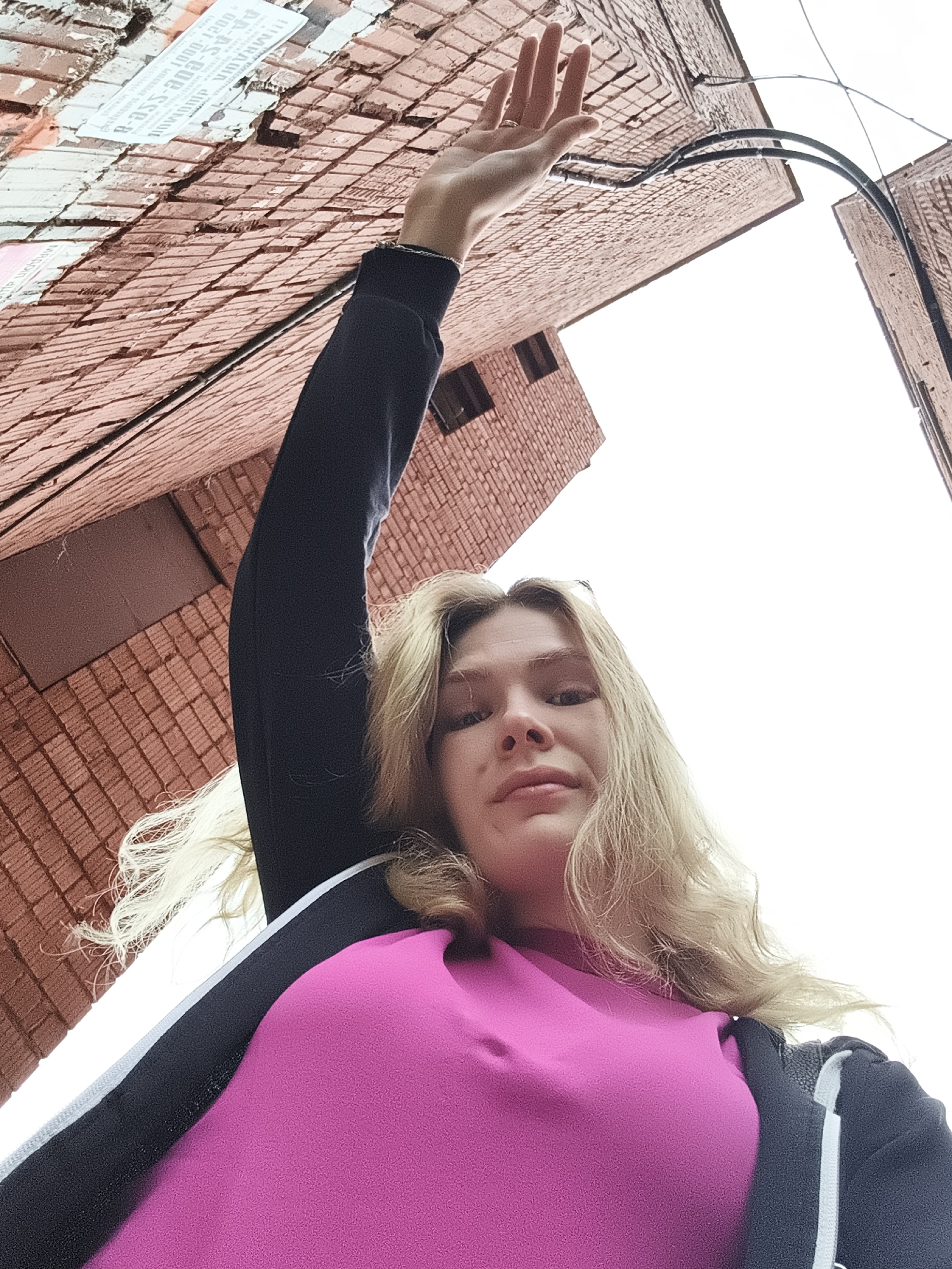 A young, fair-skinned woman with blonde hair is angled upward in the image. She is wearing a pink top and a dark jacket with a white stripe. Her left arm is raised towards the upper left corner of the image. Buildings made of red bricks are visible in the background. Electrical wires also appear in the upper right. The location is Izhevsk, Russia.
