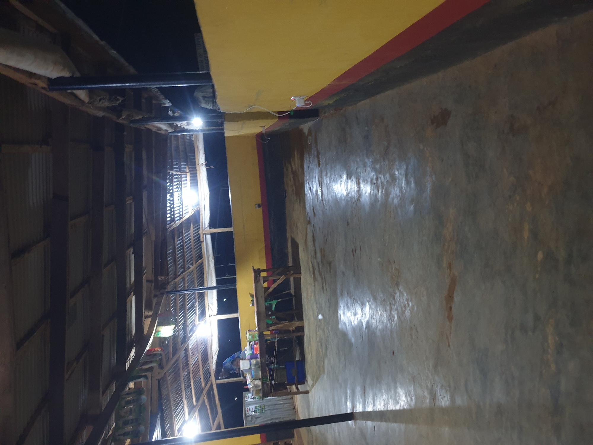 The image depicts an outdoor covered area at night. The scene appears to be a business or gathering space in Anilelerin, Nigeria. A corrugated metal roof is supported by wooden beams and metal poles. The floor is concrete and appears wet in some areas. To the left, there is a wooden table laden with various items, including bottles and containers, with a person standing behind it. A corrugated metal structure is visible behind the table. The wall on the right side of the image is painted yellow with a red stripe near the bottom. Electrical wires and a power outlet are visible on the yellow wall. The background is dark, indicating nighttime.