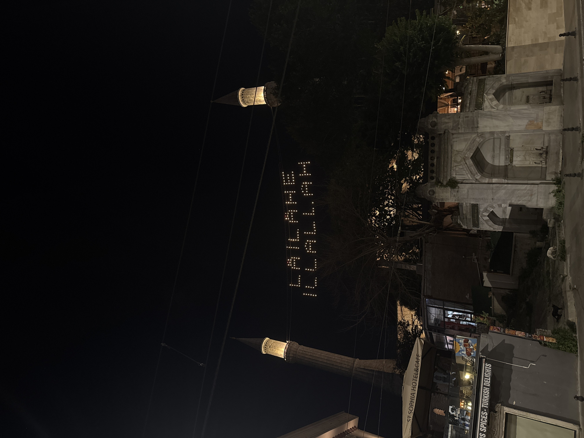 The image captures a night scene in Istanbul, Turkey, likely near Hagia Sophia due to the presence of a minaret and a building labeled "ST. SOPHIA HOTEL&CAFE". In the background, two illuminated minarets of a mosque rise against the dark sky. Stretched between them is a banner of lights forming Arabic calligraphy that reads "LAILAHE ILLALLAH" (There is no god but Allah). In the foreground, a building with a cafe awning is visible, along with a stone fountain and several cats on the street. The environment suggests a public area, possibly a street or plaza, at nighttime.