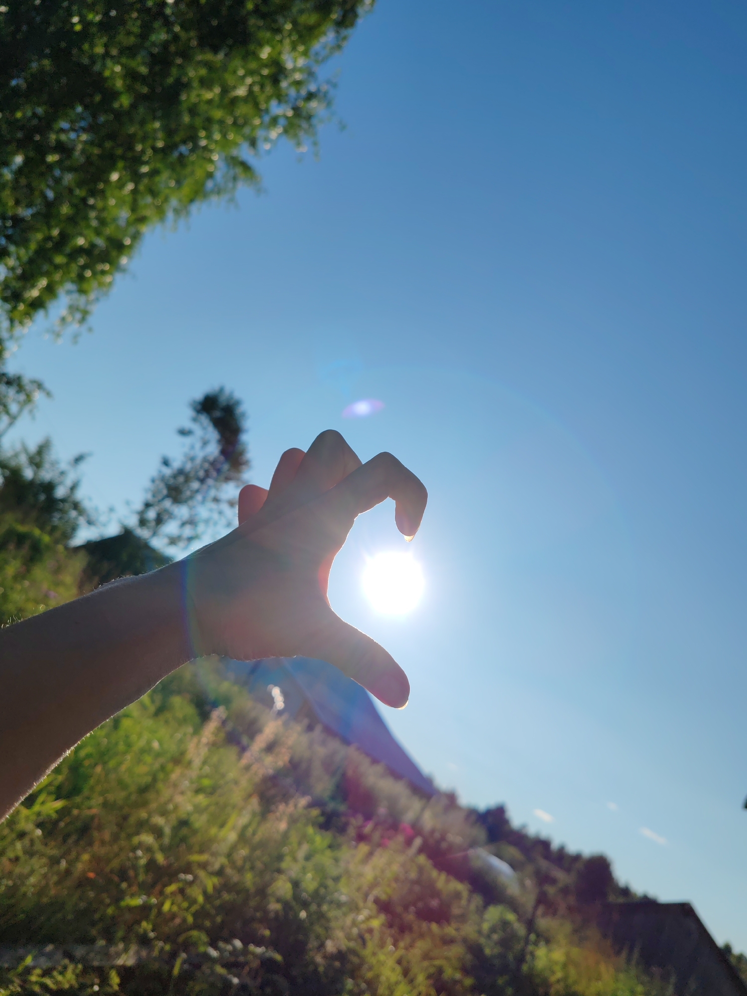 A person's hand is held up against a bright sun, forming a heart shape. The sun is positioned in the center of the heart formed by the fingers and thumb. The background is a clear blue sky with some greenery and a sloped hill or mountain in the distance. There are lens flares present in the image due to the direct sunlight. The image was taken in Чухлэм, Russia.