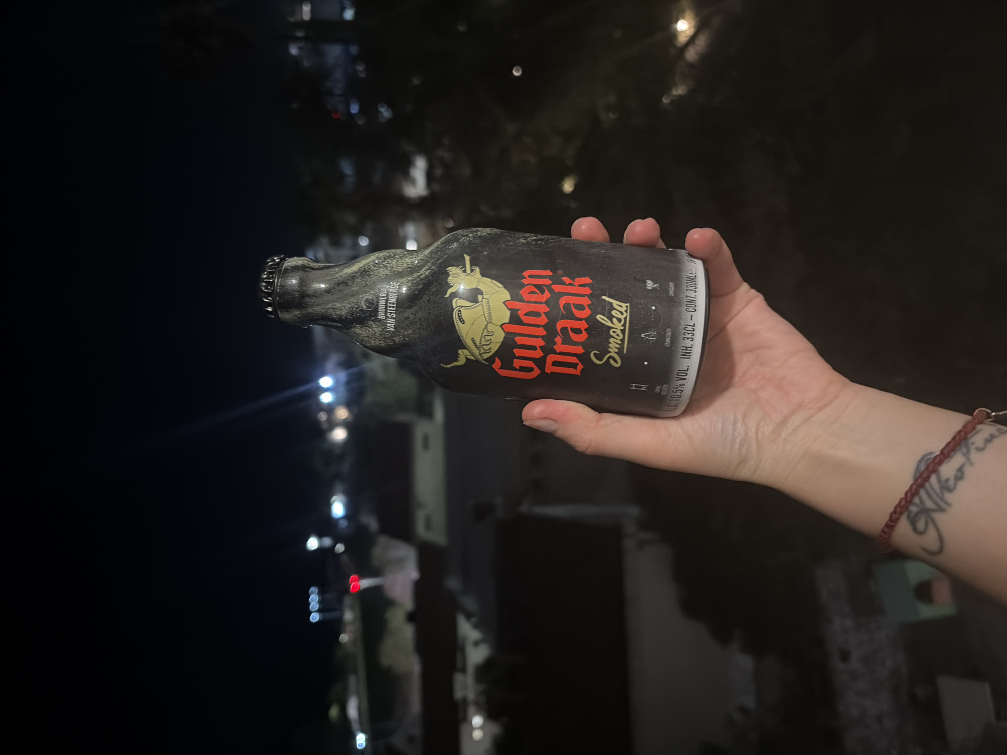 A hand holds a bottle of Gulden Draak Smoked beer, the black bottle covered in condensation. A bracelet and tattoo are visible on the wrist. Out of focus in the background are city lights, and structures that seem to indicate the location is the city of Budva, Montenegro.