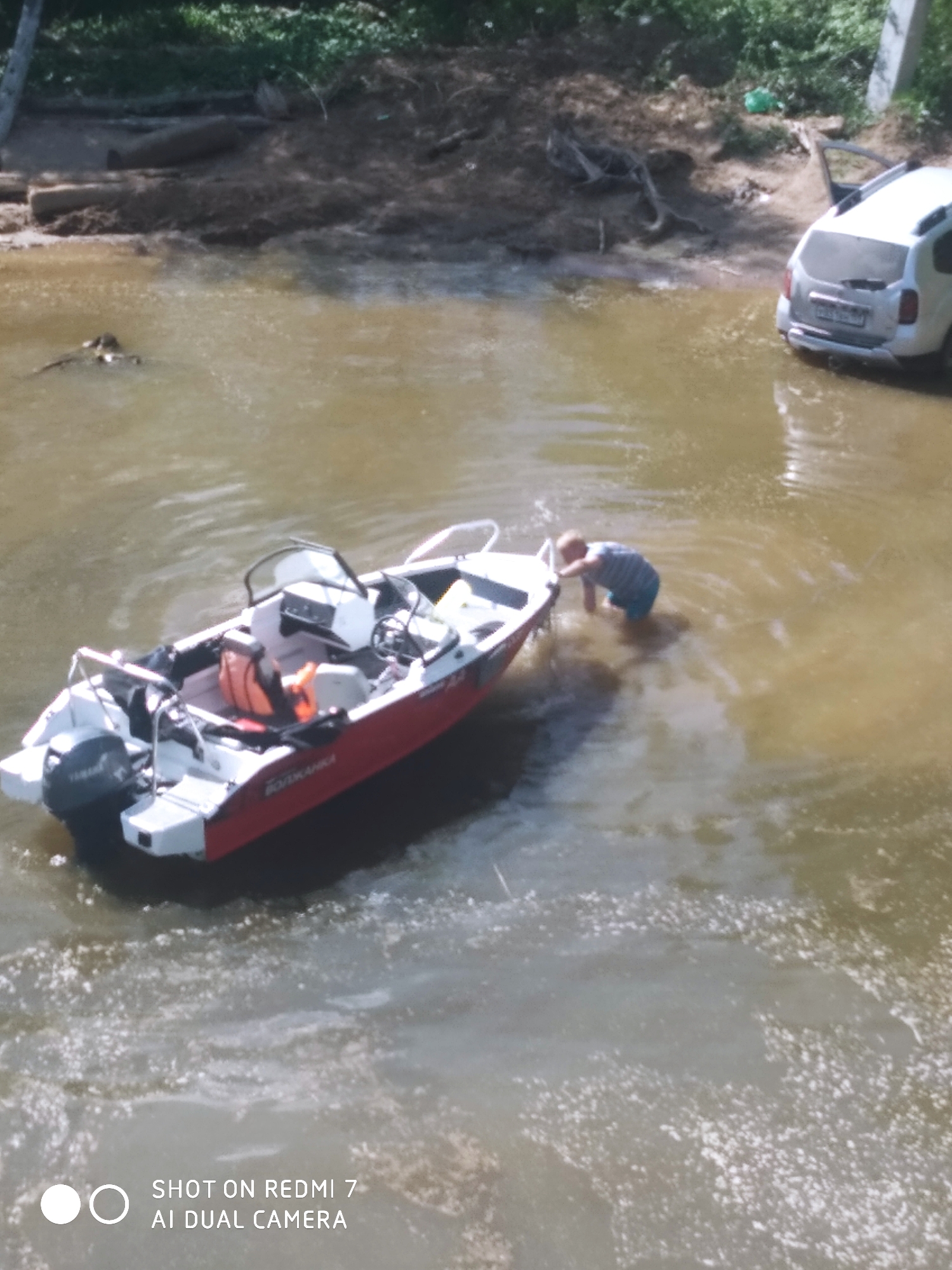 The image shows a body of water in Chaikovsky, Russia. A man is standing in the water, near a red and white boat. A white SUV is parked partially submerged on the shore. The water is murky. Vegetation and logs are visible on the shoreline.