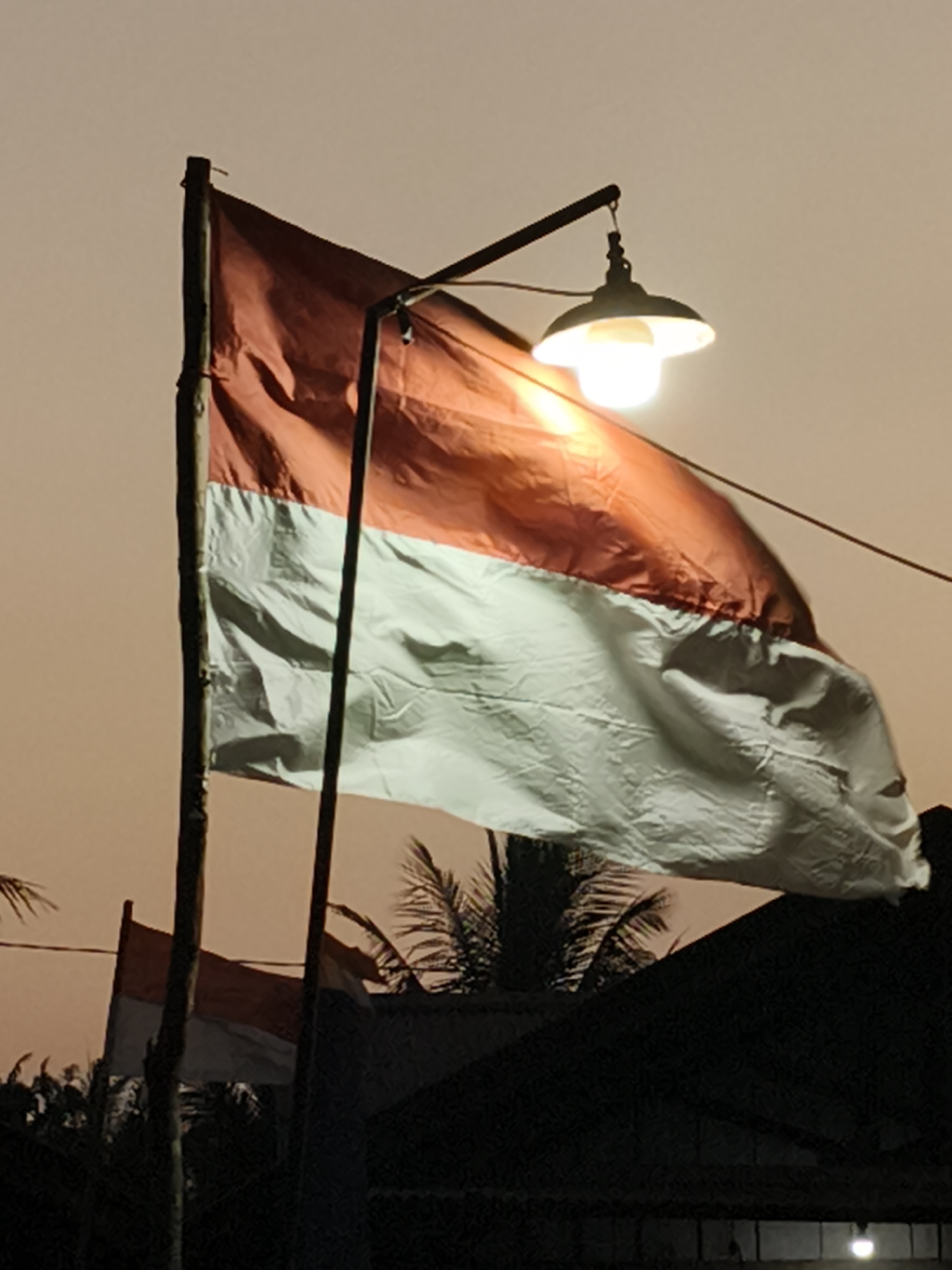 An Indonesian flag is displayed on a flagpole. A lamp hangs above the flag, illuminating it. The flag is red on top and white on the bottom. Trees and buildings are visible in the background. The sky is a dusky orange color, suggesting it is either dawn or dusk.