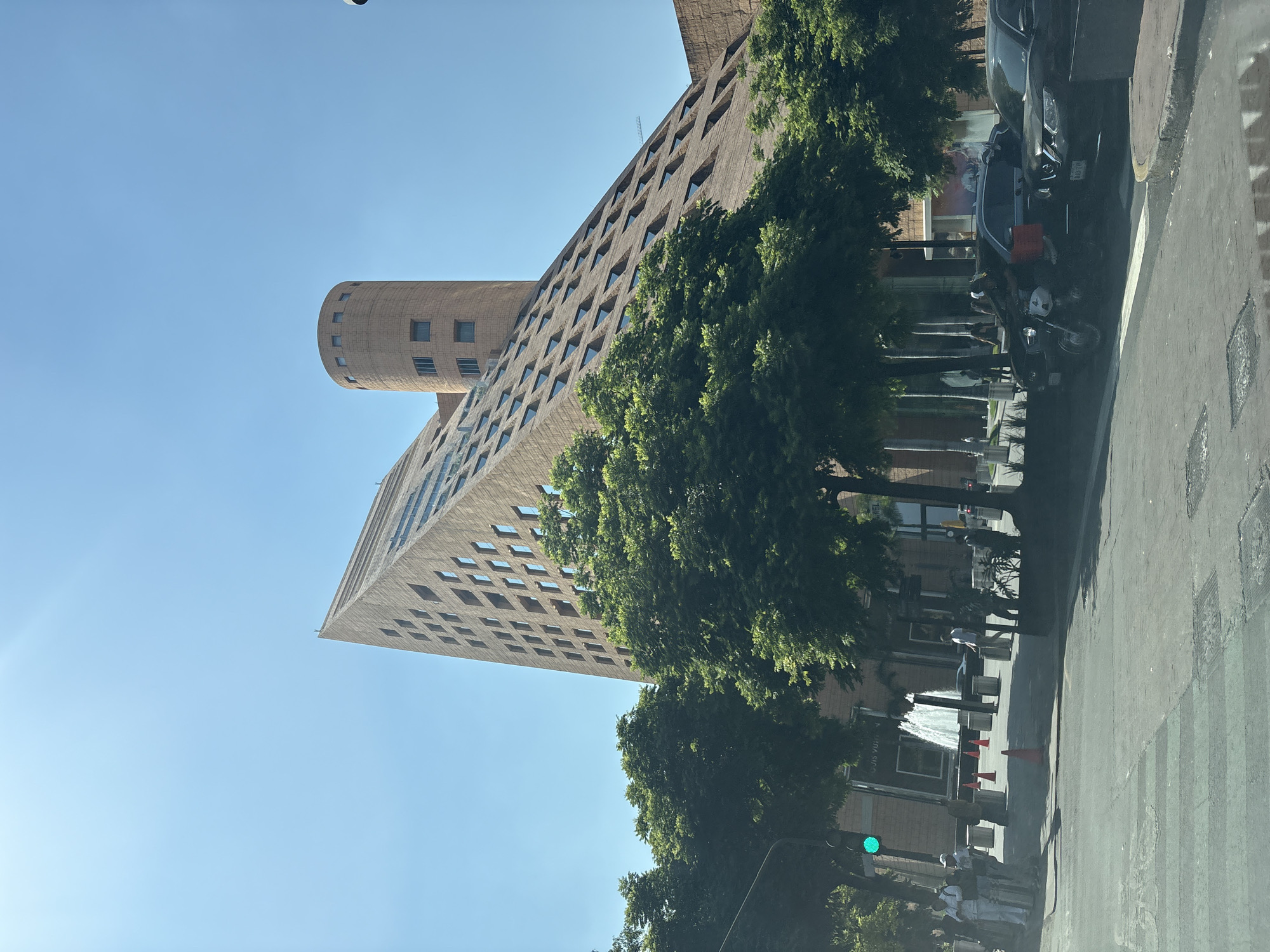 The image depicts a tall, brick building with a cylindrical tower extension, located in Mexico City, Mexico. The building has numerous small, square windows arranged in a grid-like pattern. A row of trees obscures part of the building's facade. A street runs alongside the building, where cars, motorcycles, and pedestrians are visible. A fountain is also present in the foreground. The sky is clear and blue.