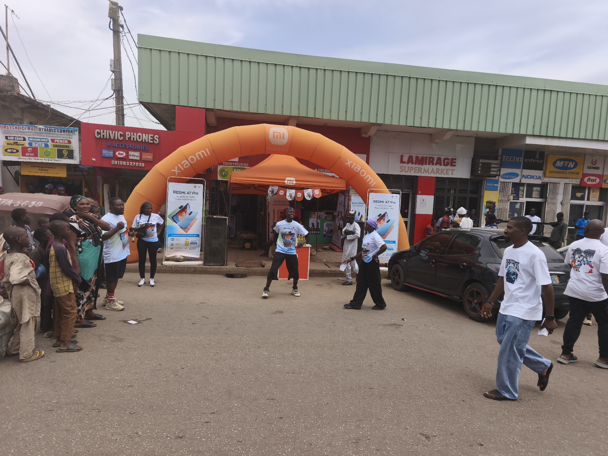 This image captures a vibrant promotional event for Xiaomi mobile phones taking place on a street in Jos, Nigeria, during daytime with an overcast sky.
The central focus is a large orange inflatable arch displaying "Xiaomi" branding and its logo, positioned over an orange canopy tent that also features the brand. On either side of the arch, large vertical display stands showcase the "REDMI A7 Pro" smartphone, highlighting its features.
Numerous people are present, including adults and children. Several individuals, likely promoters, wear white T-shirts. One man in a white T-shirt with a colorful graphic and black shorts is actively dancing in the middle of the street, engaging the crowd. Another person in similar attire stands nearby. On the left, a group of children and adults observe the activity. A man in a white T-shirt labeled "OFFICERS" and light blue jeans walks on the right, past a black car.
The event is set against a backdrop of various shop fronts. On the left, "FIRST CHOICE MULTIDYNAMIC COMPANY" advertises "SIM CARD REGISTRATION" for "MTN", "Airtel", and "9mobile", alongside "WELCOME BACK", "DATA", "POUCH", and "CHARGERS". Next to it, "CHIVIC PHONES & ACCESSORIES" prominently displays logos for "Tecno", "itel", "Infinix", and "GIONEE", with a contact number (08185327030). Further right, "LAMIRAGE SUPERMARKET" is visible, and another shop features logos for "TECNO", "Infinix", "SAMSUNG", "NOKIA", "MTN EXPERIENCE CENTRE", "GIONEE", and "itel". A utility pole with wires is also visible overhead. Several phone numbers are displayed on various signs.