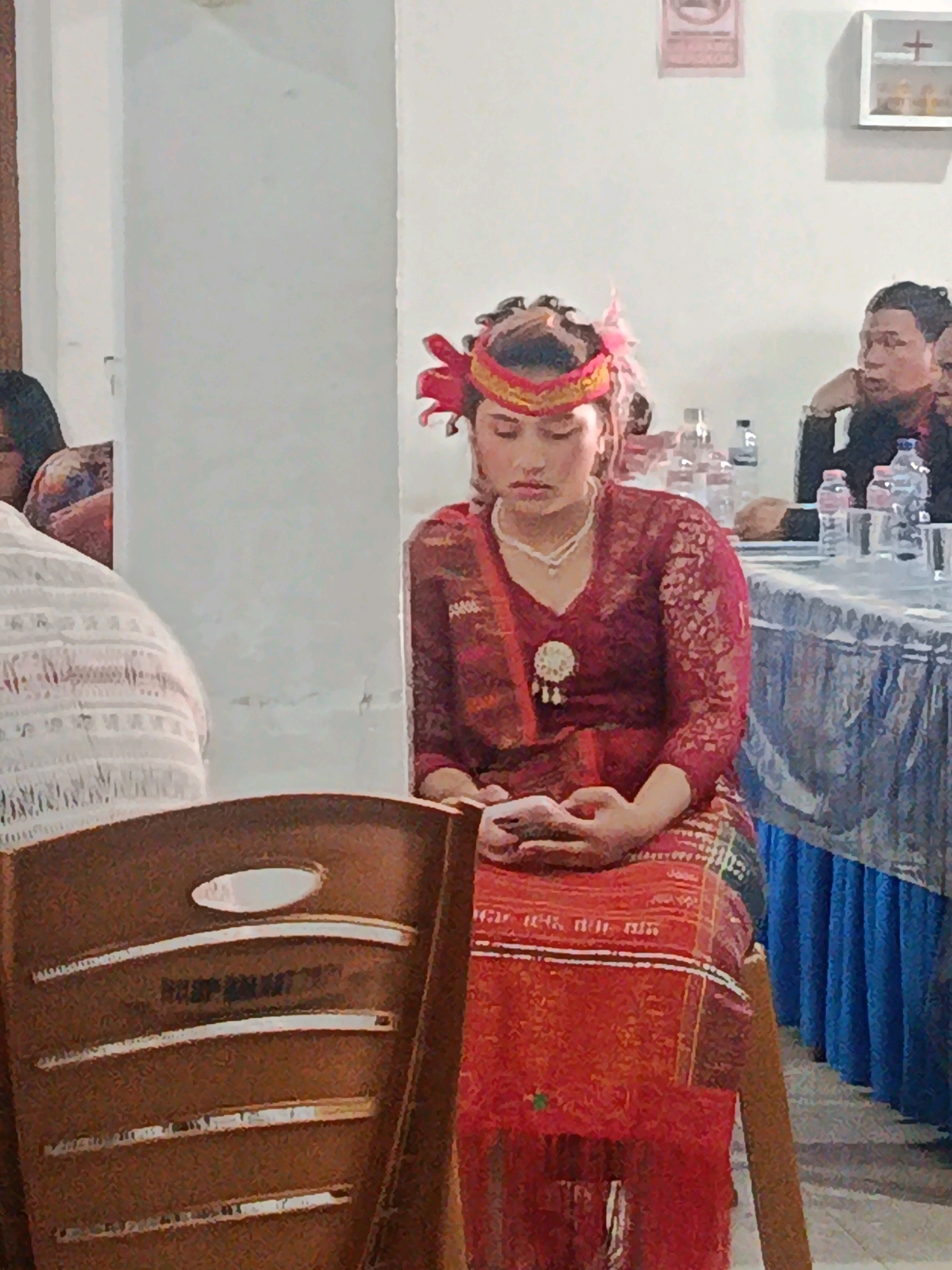 In Babelan, Indonesia, a young woman dressed in traditional red attire with intricate gold embroidery and a decorative headdress sits at a table, her hands clasped as she looks down, holding a small object. She wears a pearl necklace and a circular brooch. Behind her, another person, also dressed in traditional clothing, is partially visible. To her right, a man in a black shirt leans forward, resting his chin on his hand as he looks towards the woman. Other individuals are present in the background, and bottles of water and cups are arranged on the table. The scene appears to be an indoor event, possibly a ceremony or gathering, with a white wall and a decorated table as the backdrop.