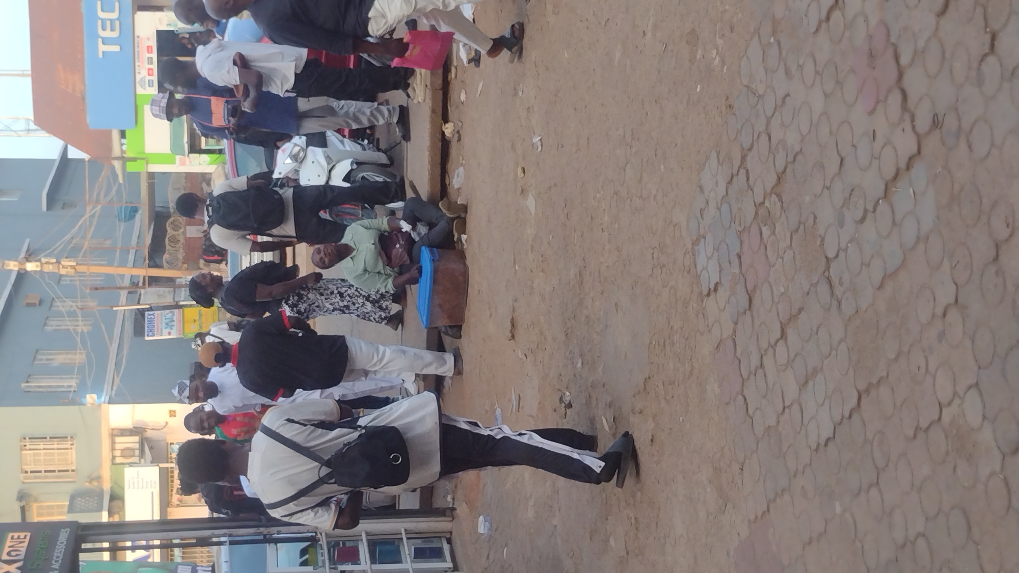 The image depicts a street scene in Jos, Nigeria. A group of people are gathered along the edge of the street near several businesses. Some individuals are standing and walking while others are seated. A man lies on the ground next to a blue cooler. Vehicles, including at least one scooter, are parked nearby. Buildings and business signage are visible in the background, including signs for TECNO and CHOMEX. The street is paved with interlocking blocks. The overall environment appears to be an active commercial area.