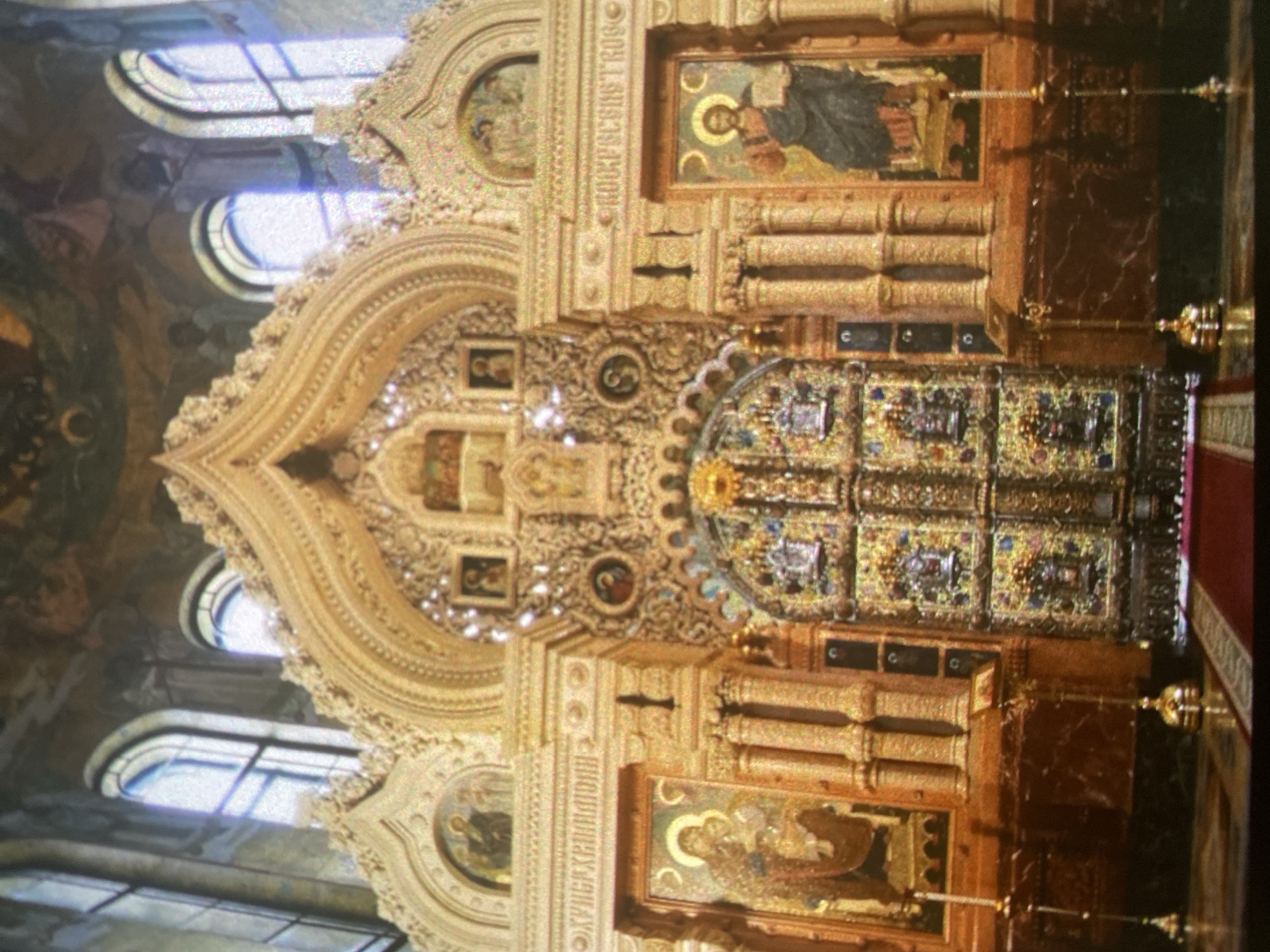 The image depicts the ornate interior of the Church of the Savior on Spilled Blood in Saint Petersburg, Russia. The focal point is the gilded iconostasis, a wall of icons and religious paintings separating the nave from the sanctuary. The iconostasis features intricate carvings, mosaics, and various religious figures. Above the iconostasis, elaborate archways and decorative elements extend towards the ceiling, adorned with additional artwork and windows. The environment is opulent and filled with religious symbolism.