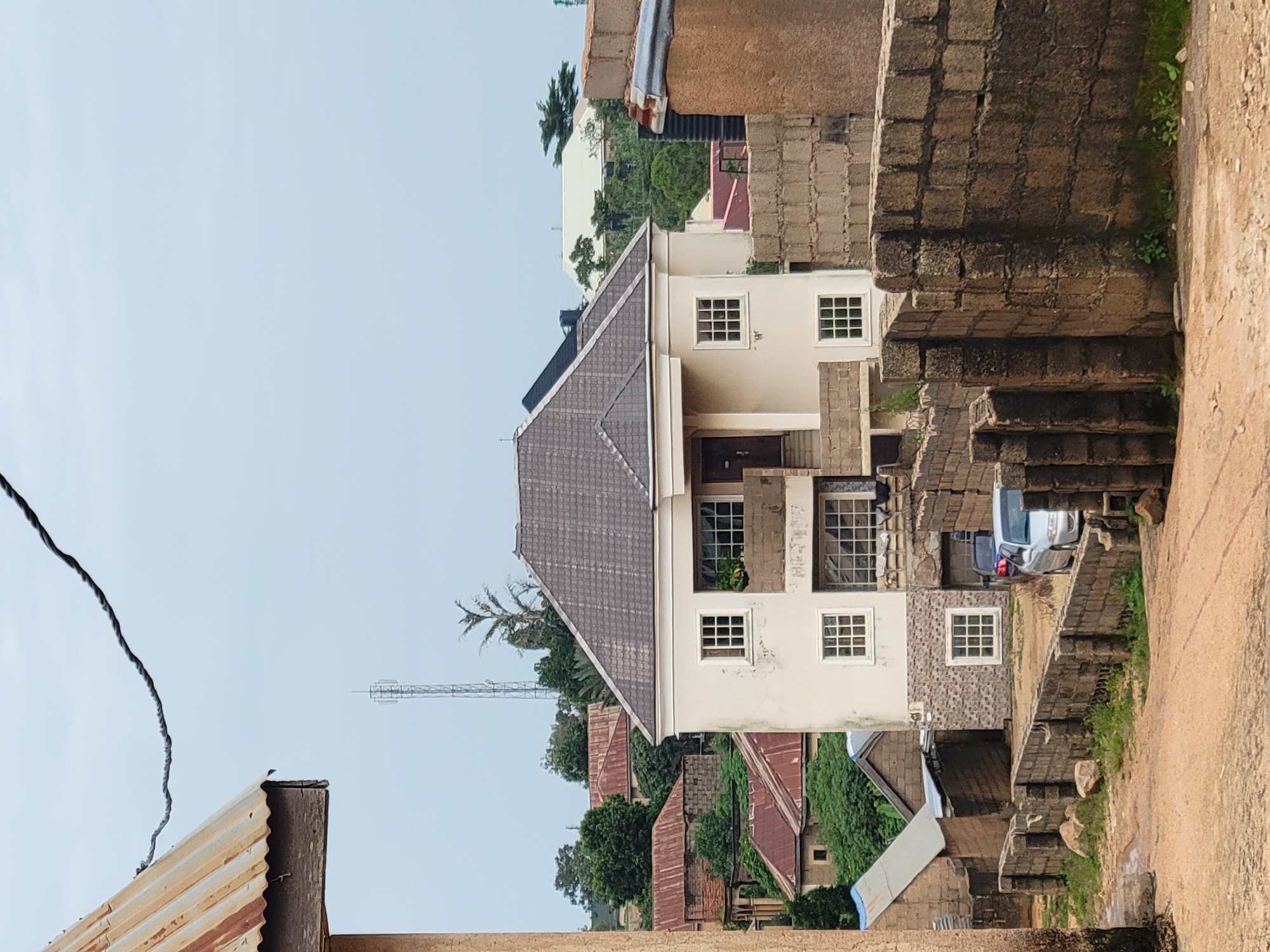 The image depicts a street scene in Jos, Nigeria, with a modern, two-story building as the central focus. This building, painted cream, features a dark-tiled hip roof, several white-framed windows, and a balcony on the upper level. In front of this house, a low wall constructed from stone blocks runs parallel to a dirt road. A silver car and a darker vehicle are parked to the right of the main house. Other buildings with corrugated metal roofs and stone block construction are visible in the background, along with lush green trees and a distant cell tower. The sky is overcast and pale. The foreground shows the dirt road with scattered rocks and some dry grass.