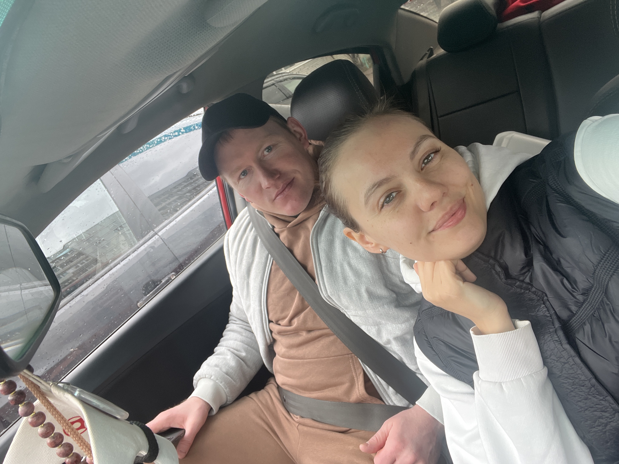 The image depicts two people inside a car, likely in Chaikovsky, Russia based on visual cues of architecture seen through the window, although this is not definitively verifiable. The man, wearing a black cap and a light brown sweatsuit underneath a grey zip-up jacket, sits in the driver's seat with his seatbelt fastened. A string of beads hangs from the rearview mirror. The woman, wearing a white hoodie underneath a black vest, is positioned to his right, seemingly taking the picture. Rain is visible on the windows.