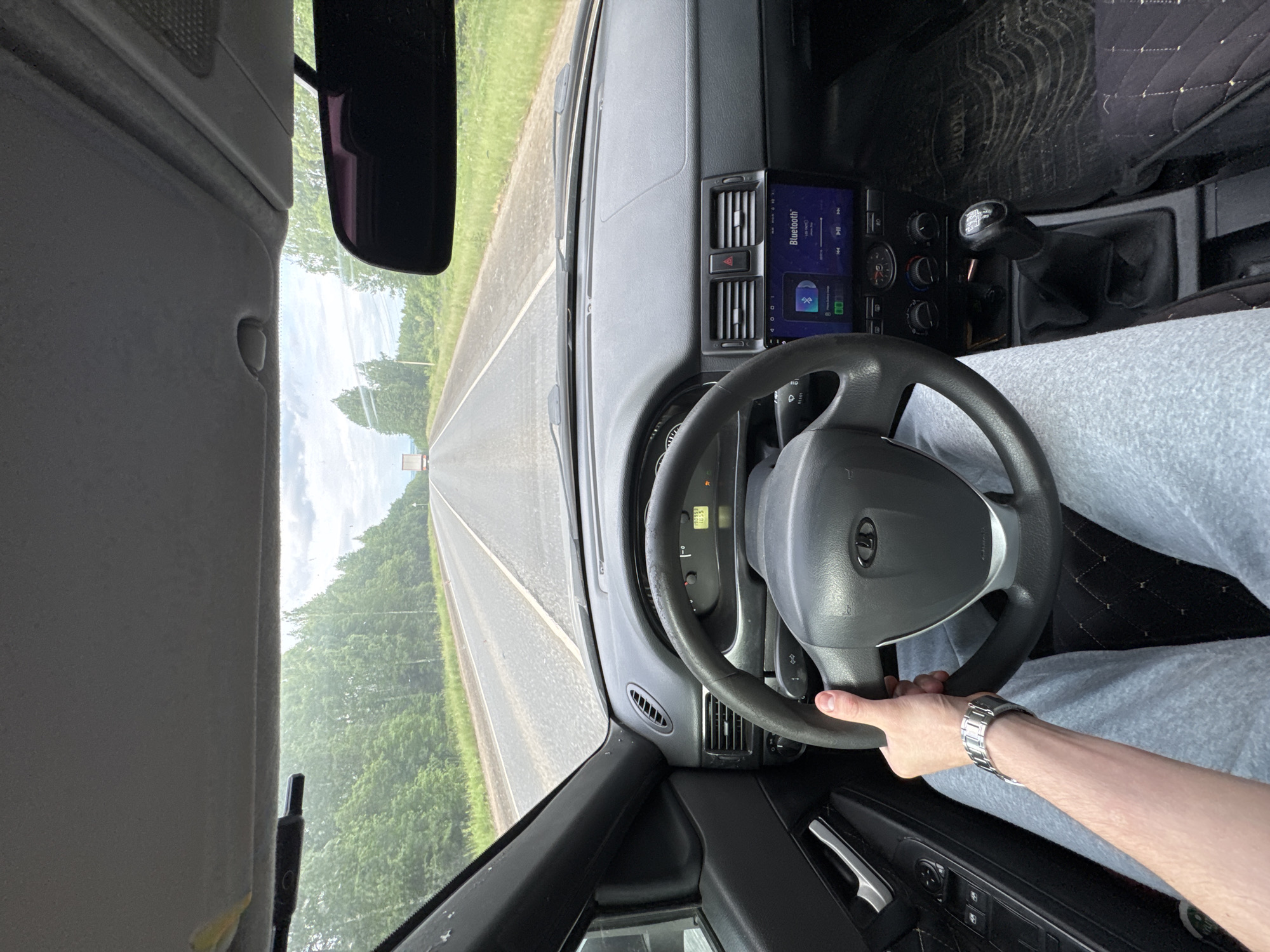 The image, rotated 90 degrees counter-clockwise, provides a driver's perspective from inside a car on a road. The scene unfolds with a paved road stretching straight into the distance, flanked on both sides by dense green forest under a cloudy sky. A faint outline of what appears to be a truck or larger vehicle is visible far ahead on the road.
Inside the vehicle, a person's right hand, adorned with a silver-colored watch, grips the steering wheel. The steering wheel prominently features the "LADA" logo in its center. The dashboard is visible, housing an aftermarket digital media unit showing a "Bluetooth" interface with music playback controls. Below this unit are various car controls, including air vents, hazard light button, and rotary knobs for climate control. A manual gear stick with unidentifiable text on its knob is located to the right of the driver. The driver's leg, clad in light-colored fabric (possibly sweatpants), is visible below the steering wheel. A patterned floor mat with "TOYOTA" text is discernible on the driver's side floor.
Other interior details include the rearview mirror, the sun visor above the windshield, and the inner door panel with window controls and a chrome handle. The car's instrument cluster is partially visible through the steering wheel, showing a digital reading of "1625". The overall impression is a casual drive during daytime under overcast conditions.
