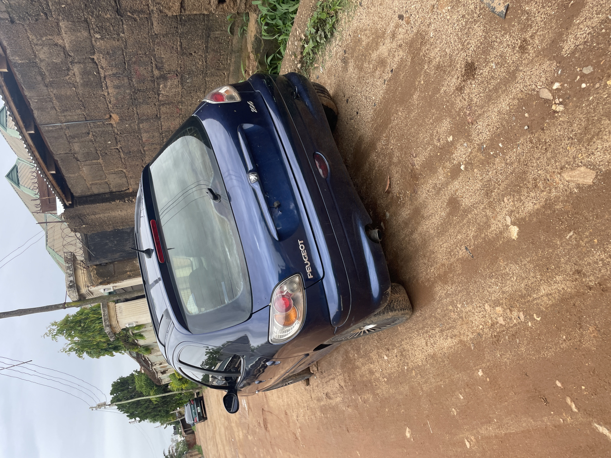 This media file captures an outdoor scene in Jos, Nigeria, featuring a dark blue Peugeot 206 hatchback parked on a dirt road. The car is angled, showing its rear and a portion of its side. The rear taillights are visible, as is the "PEUGEOT" branding on the trunk.
The setting is somewhat dilapidated, with a rough brick wall to the left and overgrown green vegetation behind the car. In the background, a building with a metal fence and barbed wire can be seen, along with electrical poles and wires, common in urban Nigerian landscapes. Further in the distance, more greenery and a cloudy sky are visible. The ground is made of dry, sandy dirt with small stones scattered about, suggesting it might be a less-maintained area or a rural part of the city. The lighting suggests it is daytime, possibly overcast due to the diffused light. No people are visible in the frame, and the overall atmosphere is neutral, focusing on the car and its surroundings. The car itself appears to be a standard model, with no unusual modifications or damage apparent from this angle, though it has a slightly worn appearance consistent with a used vehicle.