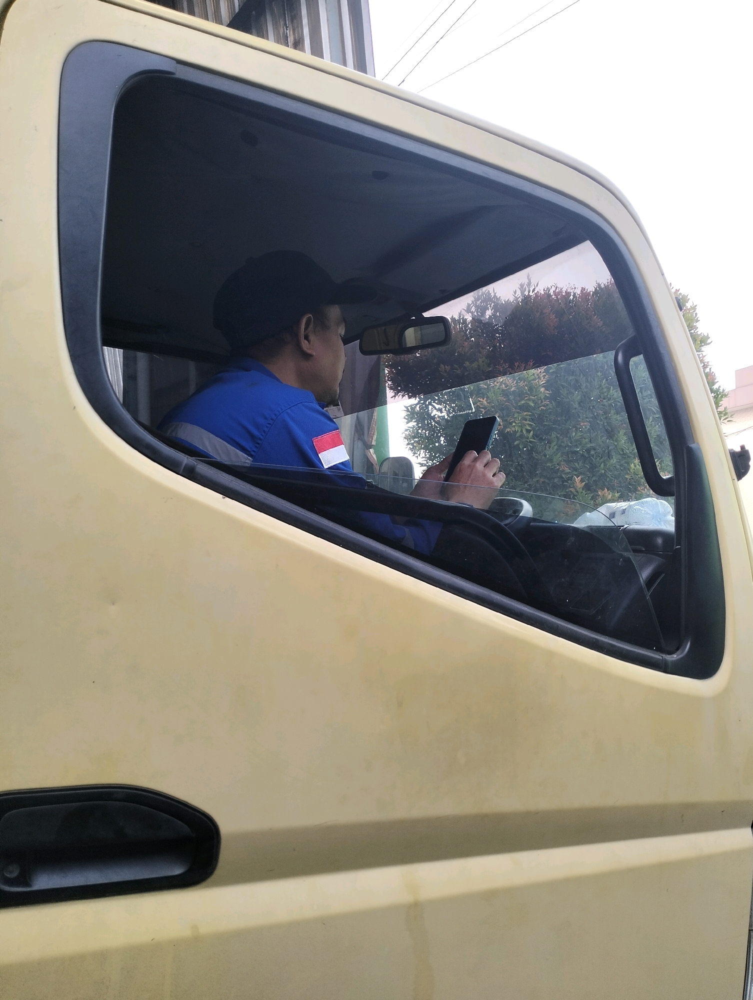 An Indonesian man wearing a blue uniform with a red and white flag patch on his sleeve and a black baseball cap is seated in the driver's side of a yellow truck, looking at his mobile phone. The truck is parked outdoors, with a blurred background of trees and a cloudy sky. The location is identified as Gunung Sindur, Indonesia.