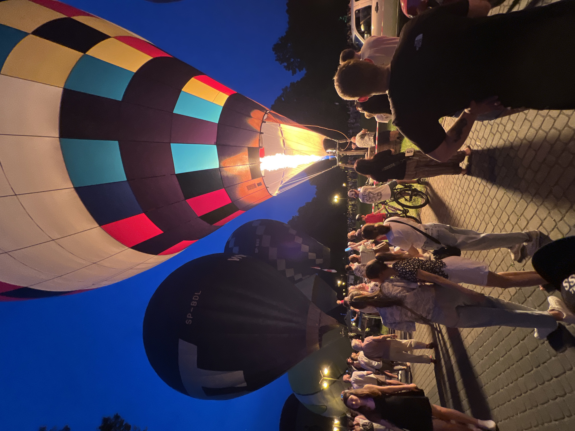 This image captures a hot air balloon festival at dusk in Tychy, Poland. The main subjects are several hot air balloons and a crowd of people gathered to watch. One large, colorful balloon is in the foreground, its fabric a vibrant patchwork of yellow, red, black, and teal. Its opening is illuminated by a powerful flame, suggesting it is being inflated. Behind it, a dark-colored balloon with the marking "SP-BDL" is visible.
The scene is set on a paved area, likely a park or public space, with a few trees and streetlights visible in the background, indicating it is nighttime or late evening. The sky is a deep blue, characteristic of twilight. The people in the crowd are diverse, with many casually dressed, suggesting a relaxed atmosphere. Some are looking up at the balloons, while others are engaged in conversation. The lighting is primarily from the hot air balloon's flame and the streetlights, casting long shadows on the pavement.
The overall mood appears to be one of excitement and anticipation, as people gather to witness the spectacle of the hot air balloons being prepared for flight. The time of day is clearly evening, transitioning into night, and the weather seems clear. The presence of a white van and bicycles suggests this might be a community event or a specific launch site. The image is oriented vertically, adding a dynamic perspective to the scene.