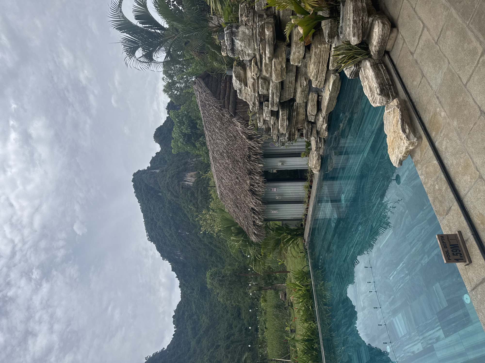 In Nam Hoa Lư Ward, Vietnam, an outdoor swimming pool is captured on a cloudy day. The pool is filled with clear blue water, reflecting the overcast sky and surrounding greenery. A small, rustic wooden hut with a thatched roof stands to the right of the pool, adjacent to a decorative rock formation. Lush vegetation, including palm trees and various plants, surrounds the area. In the background, a majestic, tree-covered mountain range dominates the landscape, with a few white string lights visible in the middle ground, suggesting an outdoor seating area. A sign near the edge of the pool indicates a depth of 1.59 meters. The scene depicts a tranquil, natural setting with resort-like amenities.