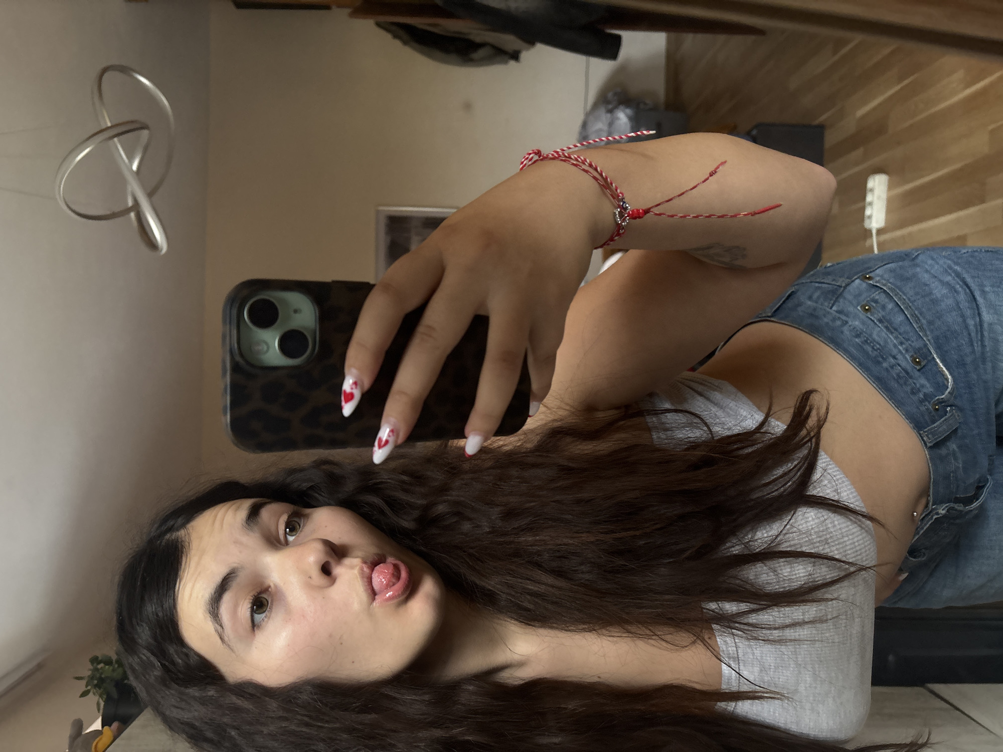 The image appears to be a selfie taken indoors by a young woman. She has dark, wavy hair, fair skin, and is wearing a gray top and blue denim shorts. Her nails are manicured with a white base and red heart designs. She is holding a smartphone with a leopard print case in her right hand, presumably taking the picture. A red and white twisted string bracelet is visible on her right wrist. Her expression is playful, sticking out her tongue.
The background shows a partially visible interior. There is a wall with what appears to be a reflection of a modern light fixture hanging from the ceiling. Other objects are discernible, including a plant and furniture, but they are out of focus. Based on the style and layout, the setting could possibly be an apartment or hotel room.
There is no definitive evidence in the image to suggest the specific location as Plovdiv, Bulgaria. Therefore, pinpointing the location in Plovdiv is not possible based on the provided image alone.
