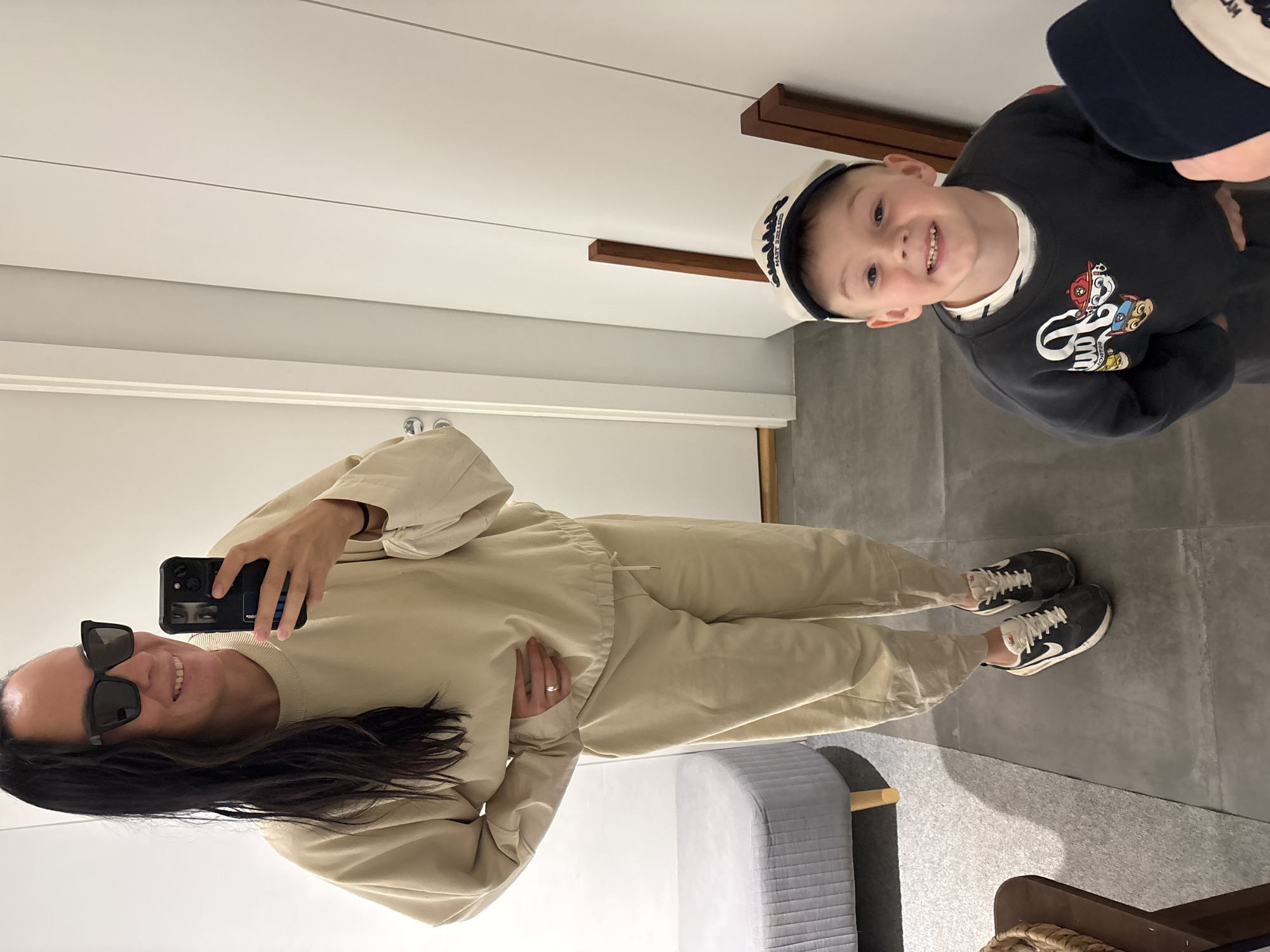 The image depicts a woman taking a selfie in a mirror with a young boy. The woman is wearing sunglasses and a beige tracksuit. The boy is wearing a navy blue sweatshirt with a cartoon print and a white sailor hat. The environment appears to be indoors, possibly a bedroom or changing room, with a bed and a grey tiled floor. No information in the photo confirms the location to be Chaikovsky, Russia.