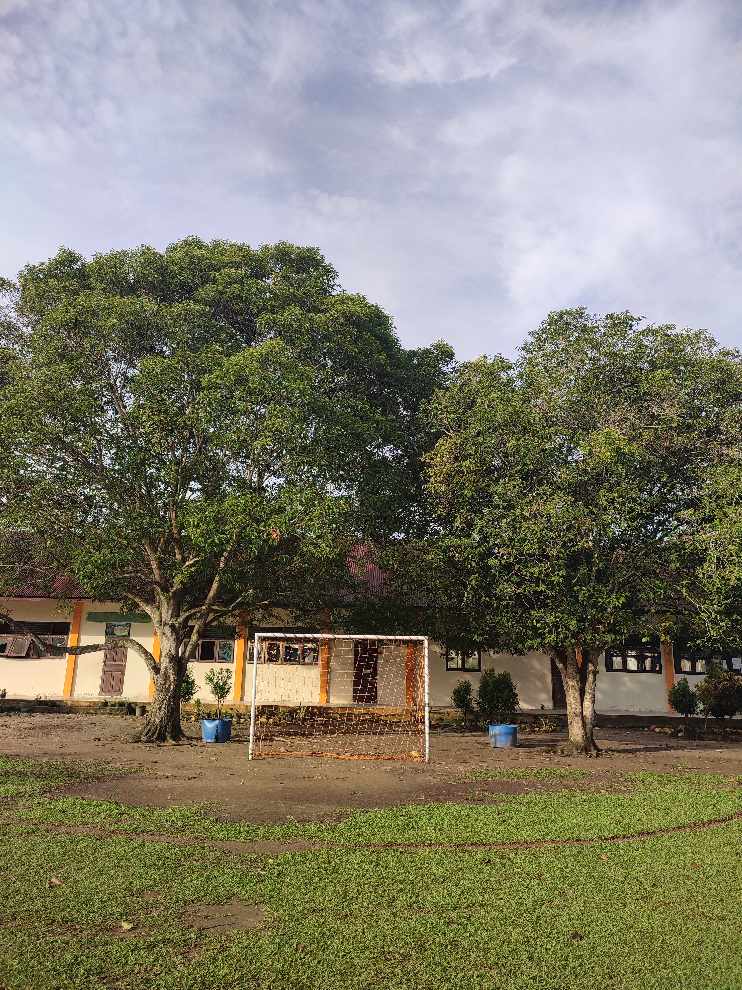 In Blangriek, Indonesia, a soccer goal stands on a dirt field in front of a school building. Two large trees with lush green foliage flank the goal, casting shadows on the ground. The school building is painted white and orange with brown window frames and doors. The sky above is partly cloudy with a mix of blue and white. The foreground shows a grassy area that appears to be part of a sports field. There are no people visible in the image.