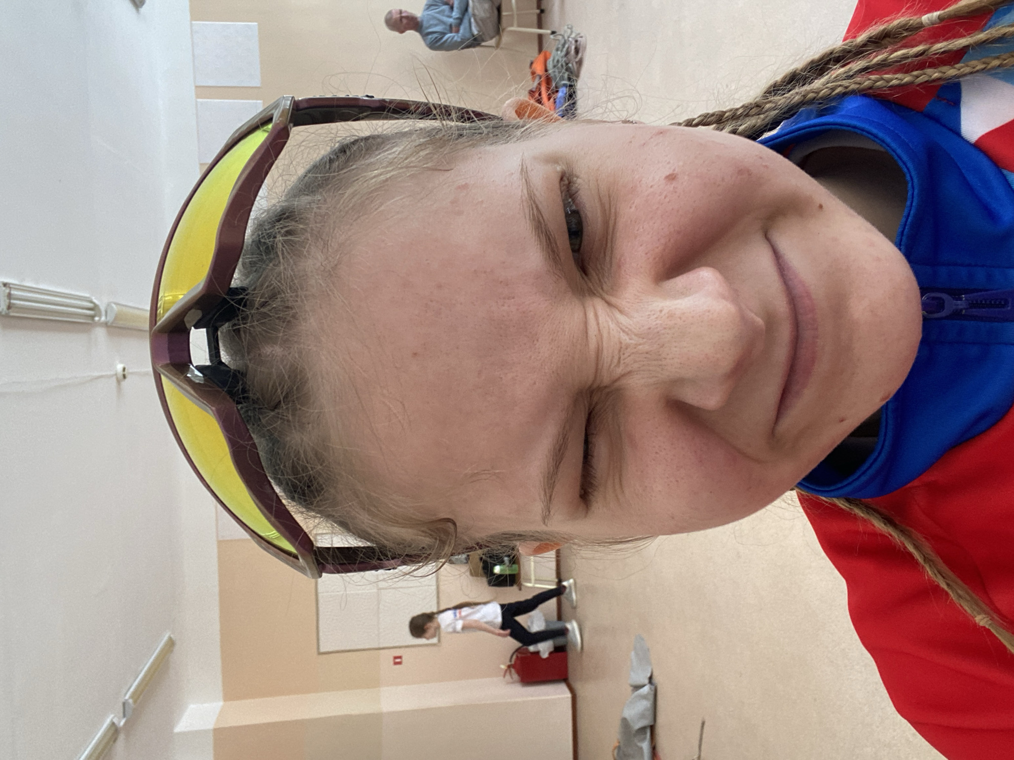 The image shows a close-up selfie of a young person with fair skin and light hair, wearing yellow-tinted sports glasses. The subject is wearing a red and blue tracksuit. The hair is styled in braids. The person is squinting and smiling slightly. In the background, other people are visible, including a child dressed in white and black clothes and one adult wearing a light-blue sweater, sitting in chairs. A red fire extinguisher is also visible. The location appears to be indoors, likely a gymnasium, with beige walls. The city of Berezniki, Russia, is not verifiable from the image.