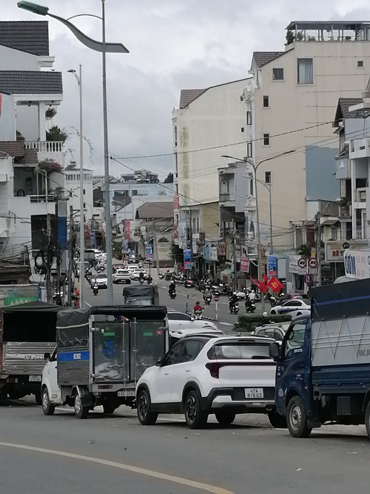 The image depicts a street scene in Da Lat, Vietnam, characterized by a mix of traffic and urban architecture under an overcast sky. The foreground features a white SUV, identified by its license plate "49A 818.63," parked alongside a blue truck with a silver cargo bed. To the left, a white truck with a black tarp-covered cargo bed is visible, with a partial license plate reading "19C 272." Behind these vehicles, other cars, motorcycles, and trucks are present, indicating moderate traffic congestion. Several red Vietnamese flags are displayed prominently on poles along the roadside.
The architecture in the background consists of multi-story buildings, typical of a city environment, with varying architectural styles, some featuring balconies and tiled roofs. Several streetlights and utility poles with numerous wires are visible, adding to the urban infrastructure. Signs in Vietnamese can be observed on the buildings, suggesting commercial establishments. The overall atmosphere appears to be a typical day in a bustling Vietnamese city.