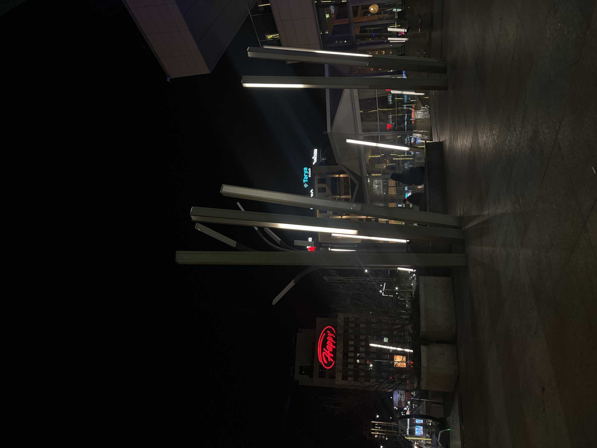 The image shows a night scene in Sofia, Bulgaria, featuring a pedestrian area with illuminated architectural light installations. Several vertical, rectangular light structures are visible. In the background, buildings display signage, including "Tarya Fintech" in teal and "Happy" in red. The ground is a dark paved surface. People are present in the background, but are difficult to make out. The sky is dark.