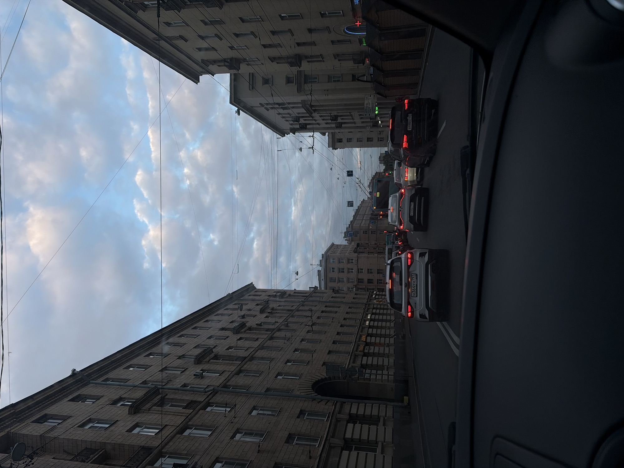 The image, taken from inside a vehicle in Saint Petersburg, Russia, captures a street scene with tall, Soviet-era apartment buildings flanking the roadway. The buildings are characterized by their uniform brick construction and repetitive window patterns. Overhead power lines stretch across the sky, which is filled with a mix of blue sky and white clouds. The street is occupied by several vehicles, including cars, in what appears to be rush-hour traffic. The perspective suggests the photograph was taken from the passenger seat. The image is taken at dusk or dawn, as suggested by the soft lighting.