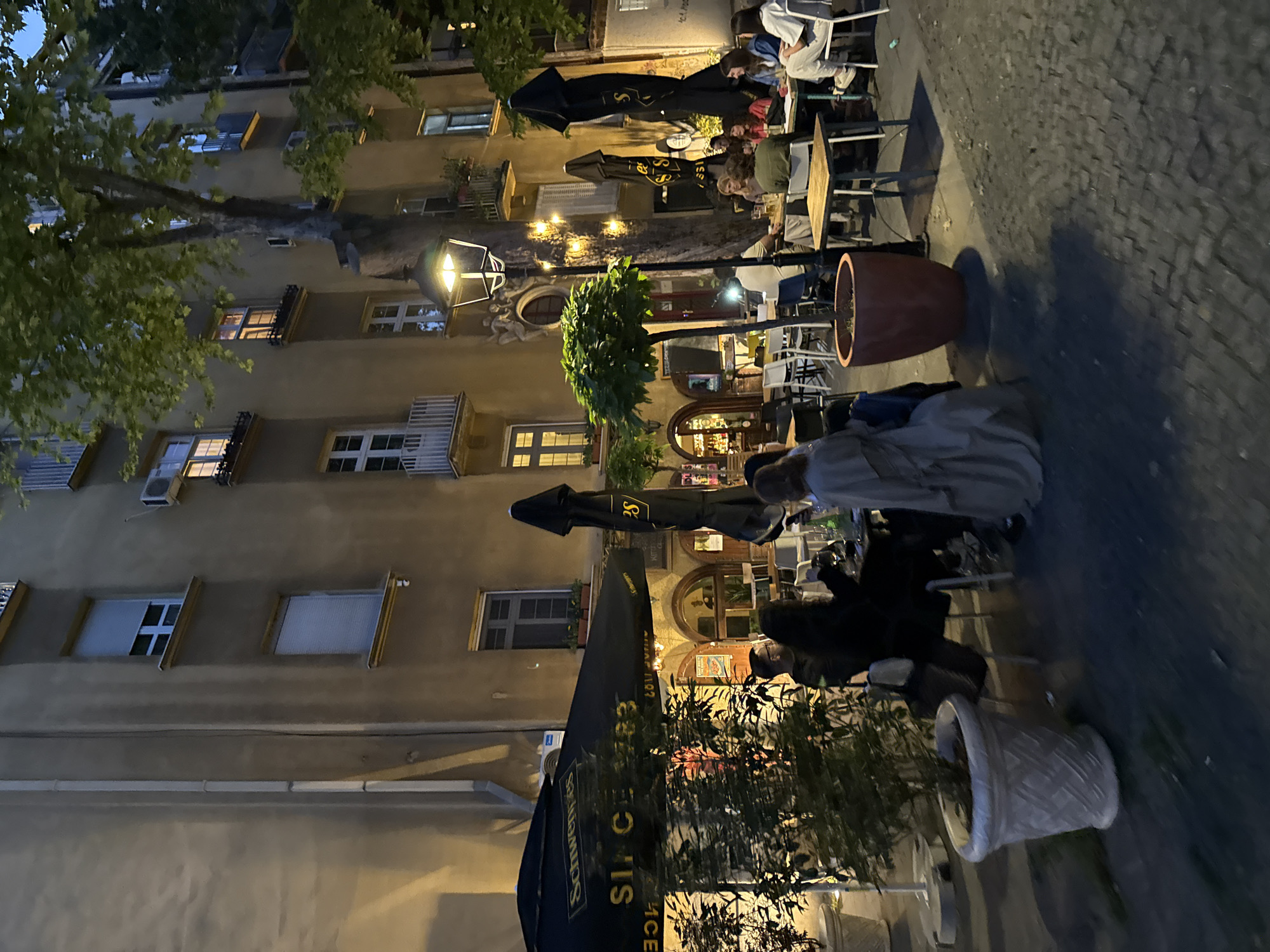 This image shows a street view of a restaurant terrace at night in Belgrade, Serbia. Several people are seated at tables, some under black umbrellas with a gold "S" logo, suggesting Schweppes branding. The restaurant is located next to a multi-story building with numerous windows, some with closed shutters or blinds, lit from within. A tree branches across the building facade, and a street lamp is visible. Several large potted plants are placed along the terrace. The ground is cobblestone.