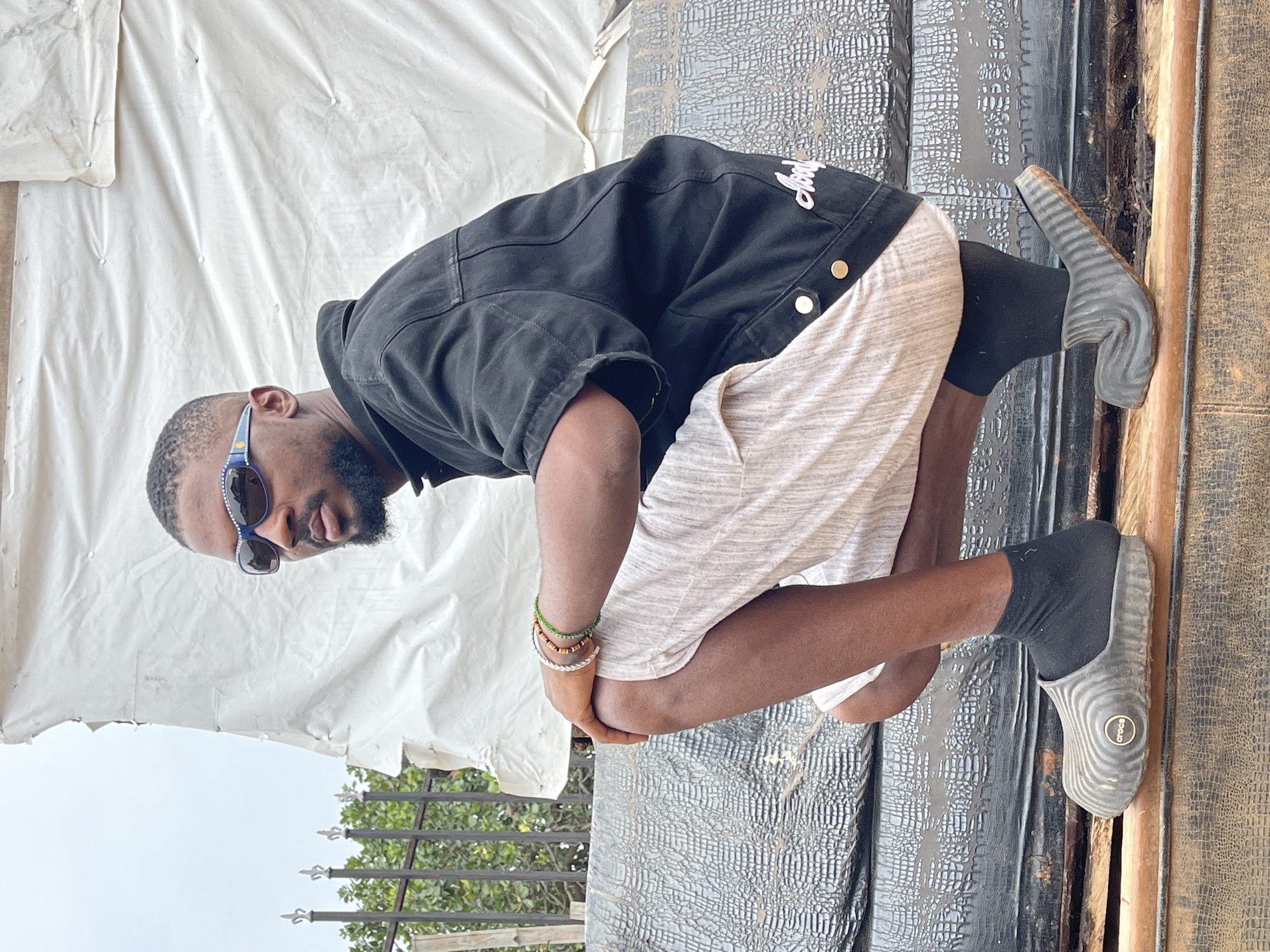 A Black male is crouching on a raised surface. He is wearing a black short-sleeved denim jacket with gold buttons, light-colored striped shorts, black socks, and gray Croc-style clogs. He has on sunglasses with blue frames and white embellishments, and several beaded bracelets on his left wrist. He is facing towards the right of the frame. Behind him, a white tarp is stretched taut and attached to poles. To the left of the tarp, a black metal fence with ornate tops is visible, with green foliage behind it. The surface he is crouching on appears to be made of dark, textured material. The overall lighting suggests an outdoor, overcast day. The location is specified as the city of Abuja, Nigeria.