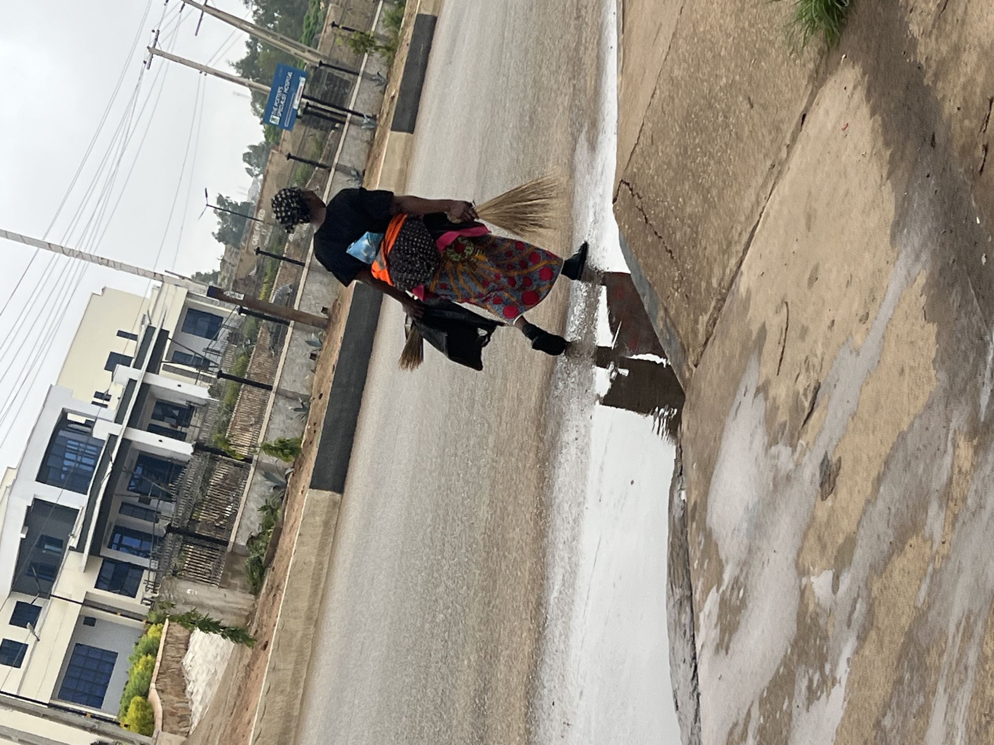 This image captures an urban street scene in Gura Top, Nigeria, on an overcast day, likely after recent rainfall. The main subject is a person, appearing to be a woman, walking along a wet, paved road next to a concrete sidewalk. She is viewed from the side, dressed in a black long-sleeved top, a vibrant, multi-colored patterned skirt or wrap, and black boots. An orange sash or vest is visible around her waist, and she wears a black and white patterned head covering. She carries a bundle of straw brooms in her right hand, held upright, and a black bag, possibly containing another broom, in her left hand.
The road and sidewalk are visibly wet with standing puddles, reflecting the grey sky, which indicates recent or ongoing rain. In the background, several modern, multi-story buildings with light-colored facades and dark-tinted windows line the street, separated by a metallic fence. Tall utility poles with a dense network of overhead power lines are prominent features, along with modern streetlights. Further in the distance, trees and other structures can be seen under the cloudy sky.
A blue rectangular sign, mounted on a pole in the background, clearly displays the text "THE POTTERS SPECIALIST HOSPITAL," providing a specific location cue. The scene depicts an everyday moment, with the individual seemingly engaged in work or commuting, set against a backdrop of developing urban infrastructure.