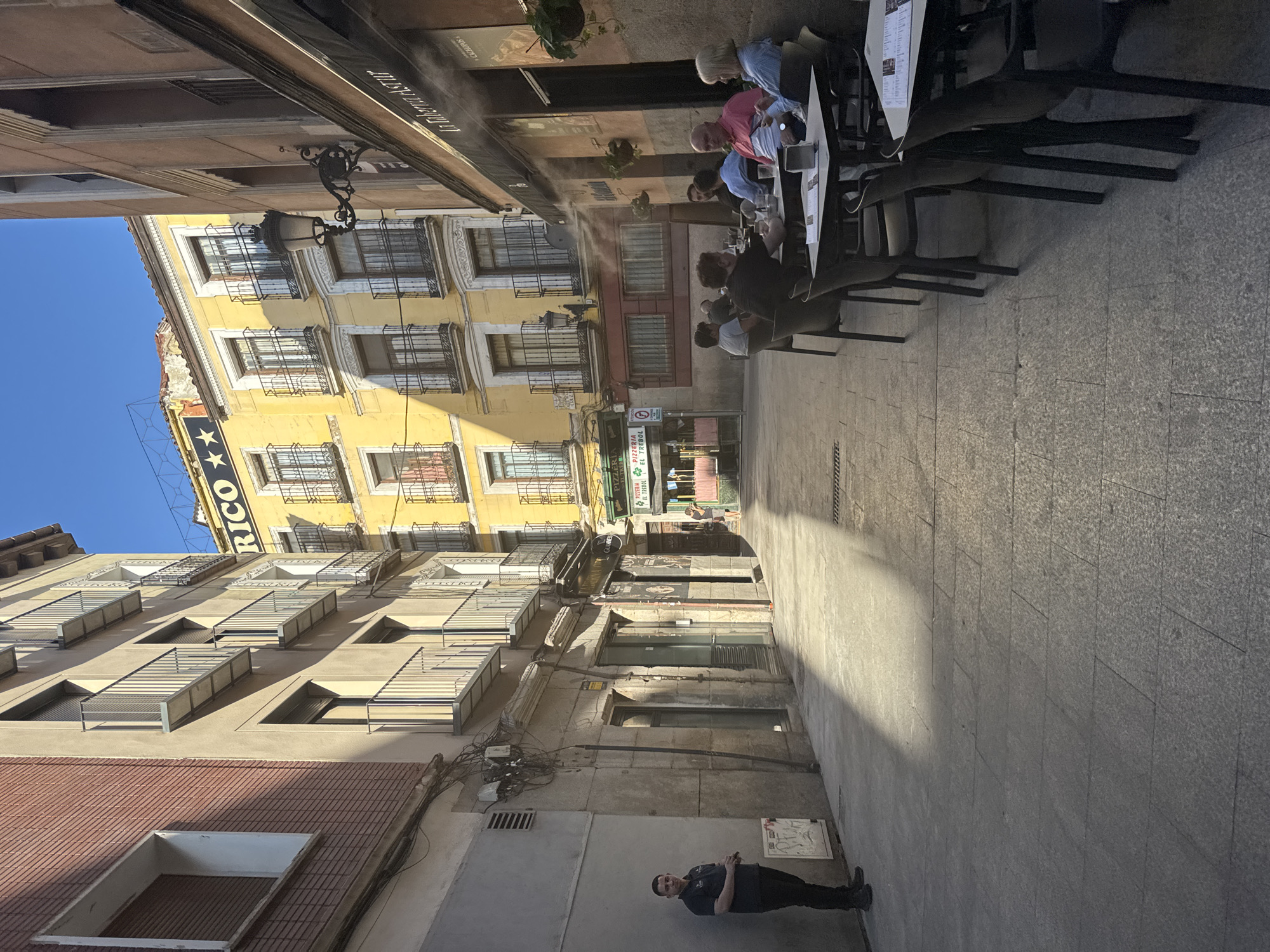 The image captures a sun-drenched street scene in Madrid, Spain, likely in a historic district given the architecture. In the foreground left, a security guard stands attentively near a textured gray wall, his dark uniform contrasting with the bright sunlight. The street itself is paved with large, grey flagstones, with a sharp line of shadow cast diagonally across it from the buildings on the right.
On the right side of the street, an outdoor cafe is in operation. Several patrons are seated at tables, some engaged in conversation, others looking at their phones or enjoying their drinks. A misting system is active, creating a visible haze of water vapor above the tables, providing a cooling effect. The cafe's facade is a warm, peachy color, adorned with a dark awning and signage, including "La Taberna Astur."
Further down the street, a vibrant yellow building dominates the view, featuring multiple balconies with railings and arched windows. A prominent sign on this building reads "RICO" with stars above it. Other establishments are visible, including a pizzeria with signage that includes "PIZZERIA EL TREBOL." The buildings exhibit classic European urban architecture with ornate balconies and window frames. The sky is a clear, bright blue, indicating a sunny day. The overall impression is one of a lively urban setting, with a mix of daily life and outdoor dining.