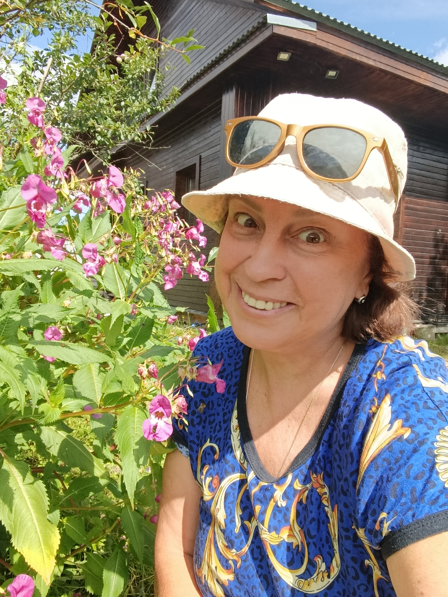 A woman wearing a light-colored bucket hat and sunglasses is taking a selfie outdoors. She is smiling, with her mouth open. She is wearing a blue t-shirt with a leopard print and a gold ornate pattern. Behind her is a wooden building with a dark roof and a large bush with pink flowers. The scene appears to be in a rural or suburban area, possibly in Vereray, Russia, given the wooden building and natural surroundings. The lighting suggests it is daytime.