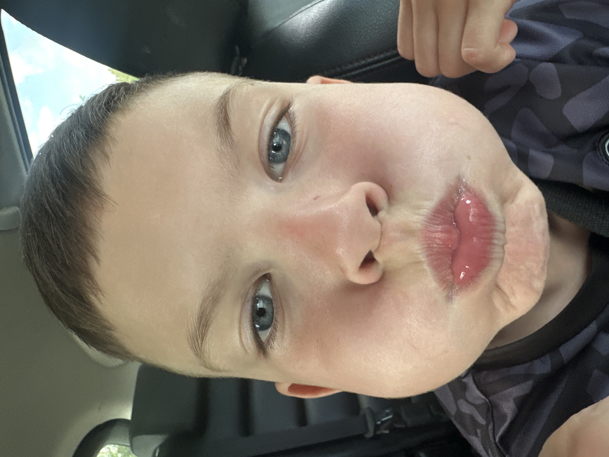 A close-up, rotated, and slightly blurry image captures a young, light-skinned child with short brown hair and blue eyes. The child is making a pucker face with their lips. The child is wearing a patterned dark shirt. The child appears to be in a car. There is no evidence to suggest the location is Chaikovsky, Russia.